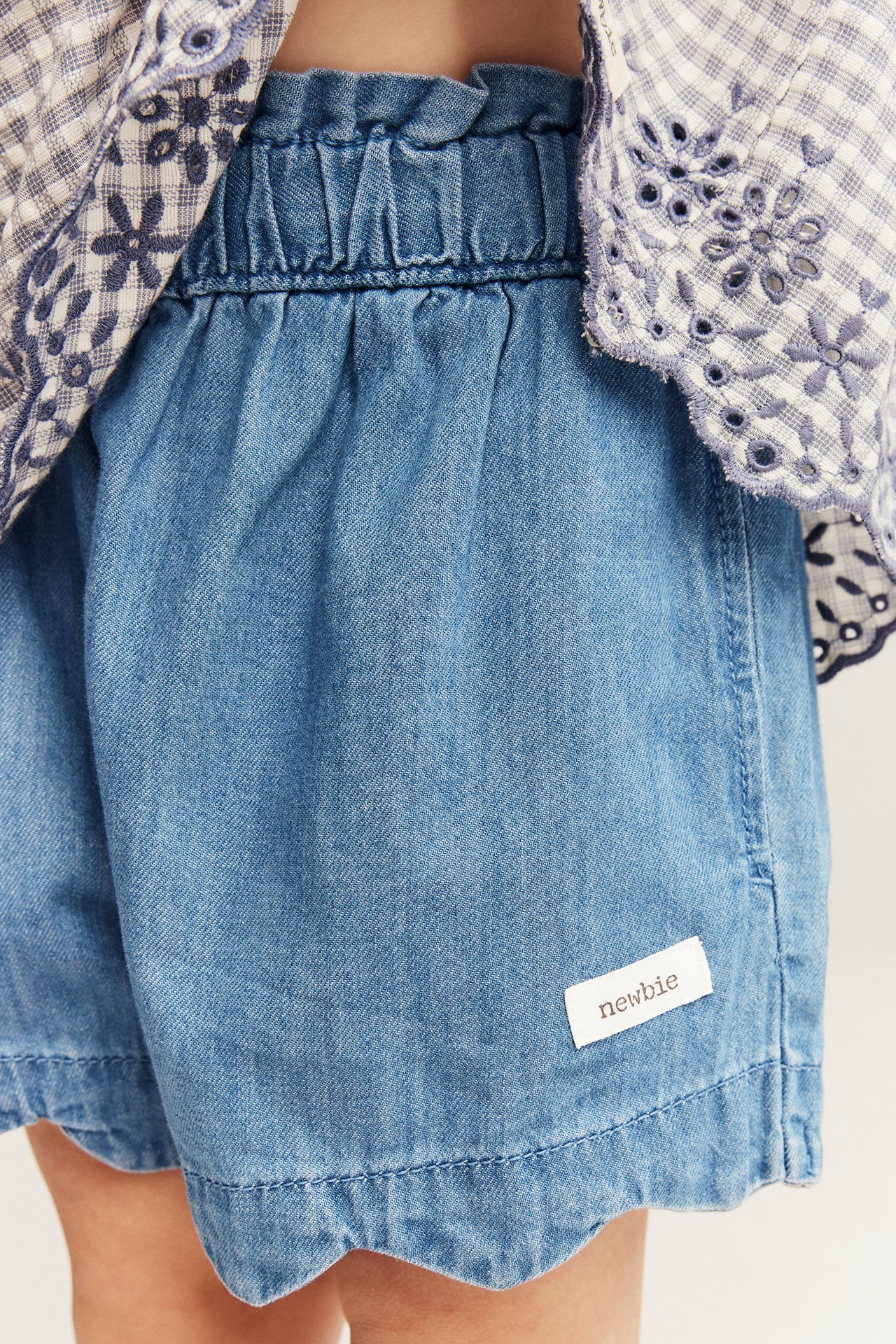 Close-up: Kid wears light blue denim shorts with ruffled waist, scalloped hem. Paired with a checkered top.