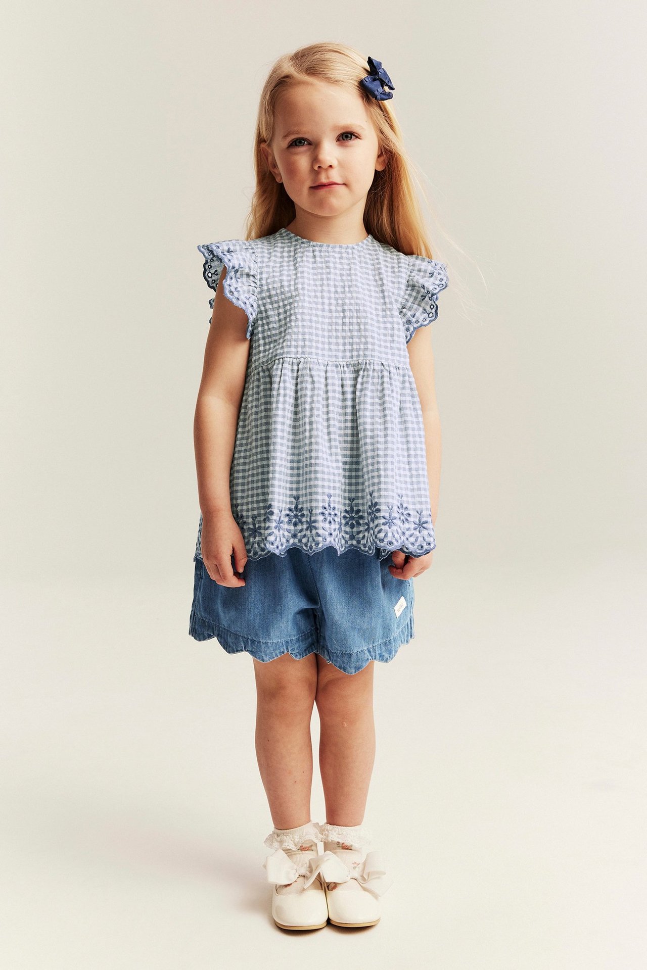 Vorderansicht: Kind in hellblauen Jeansshorts mit Wellenkante, blauem Vichy-Oberteil und cremefarbenen Schuhen.