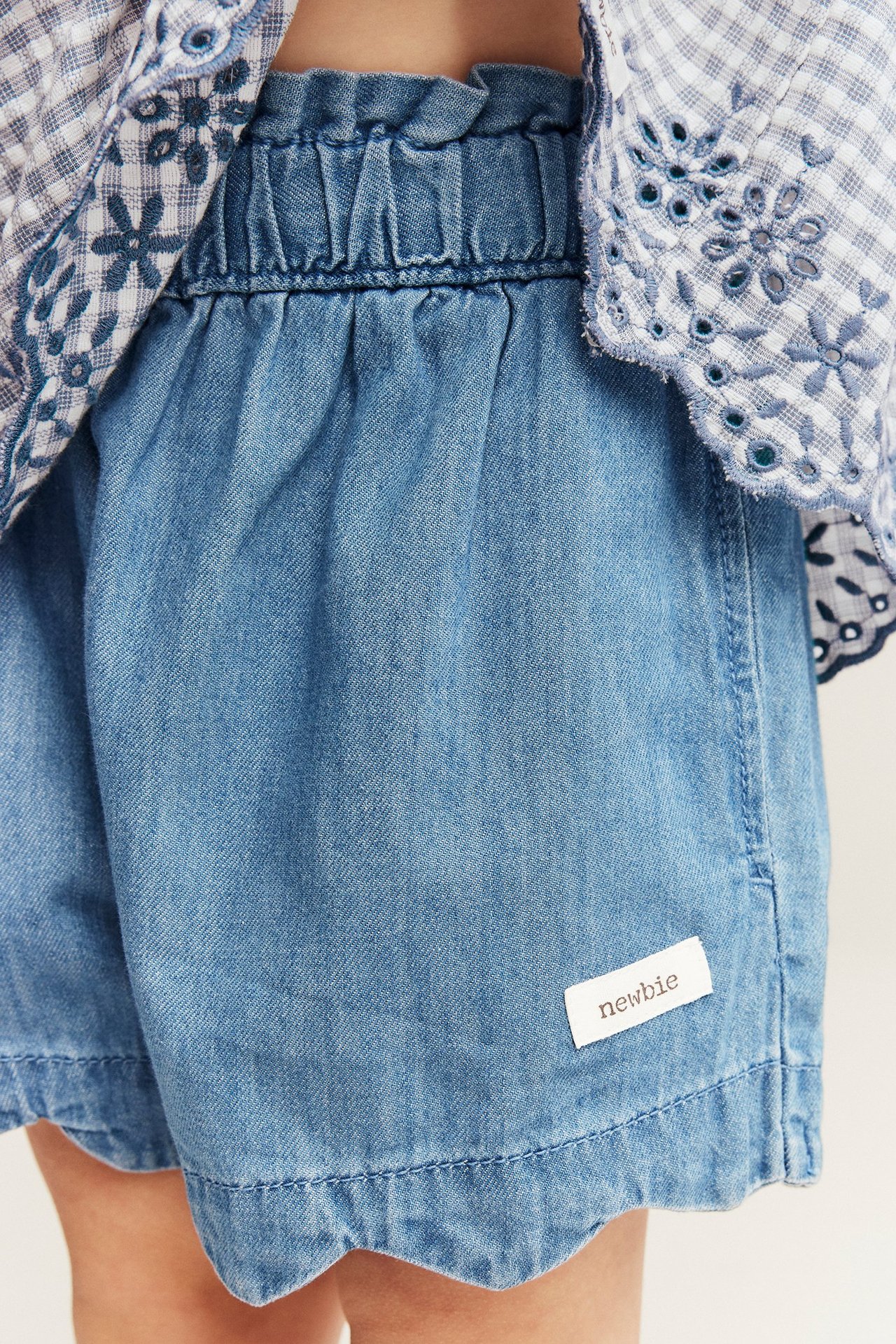 Close-up front: kid's light blue denim shorts with ruffly waist and wavy hems, with checkered shirt.