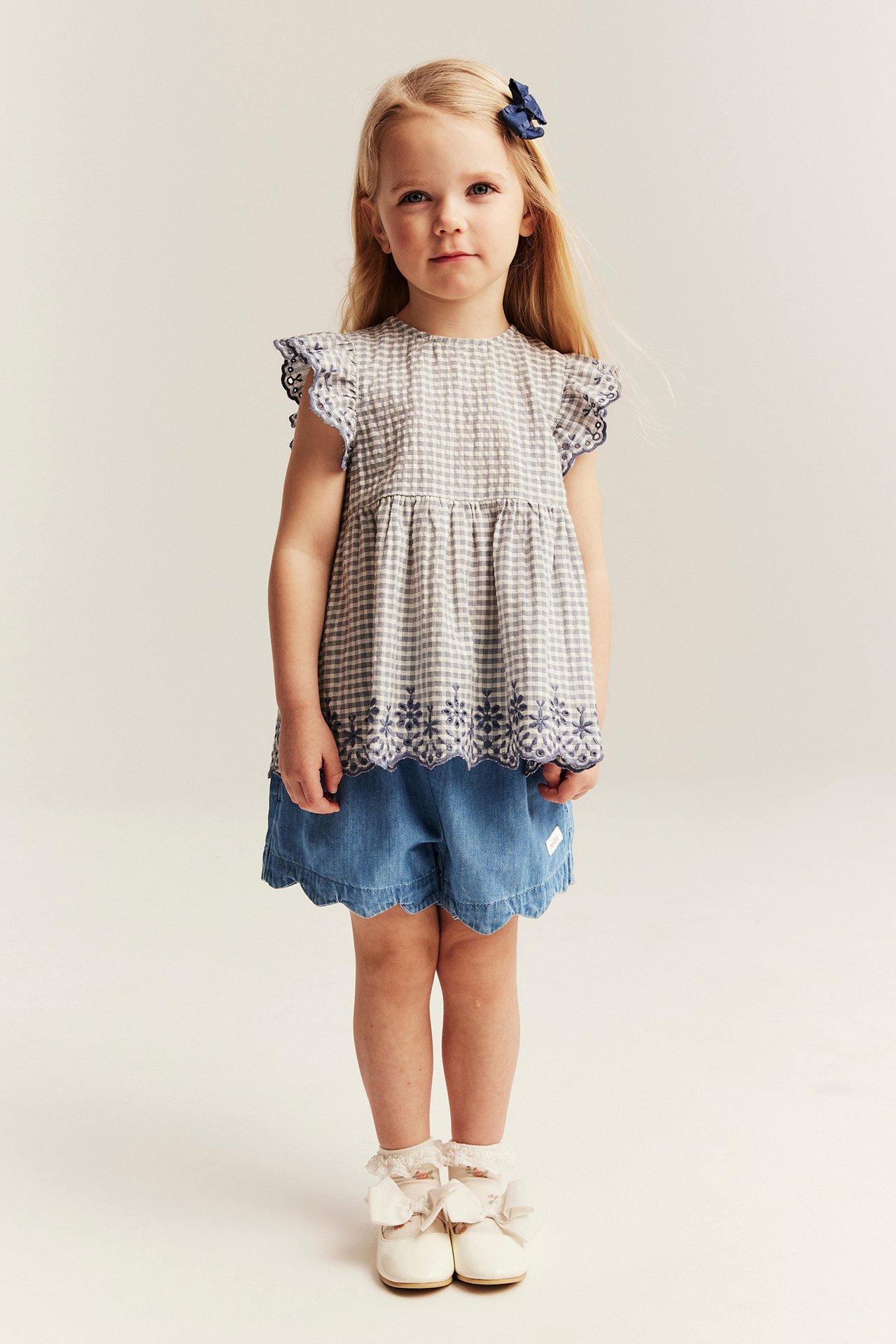 Front view: kid in light blue woven denim shorts with scalloped hem, gingham top, blue hair bow, and cream shoes.