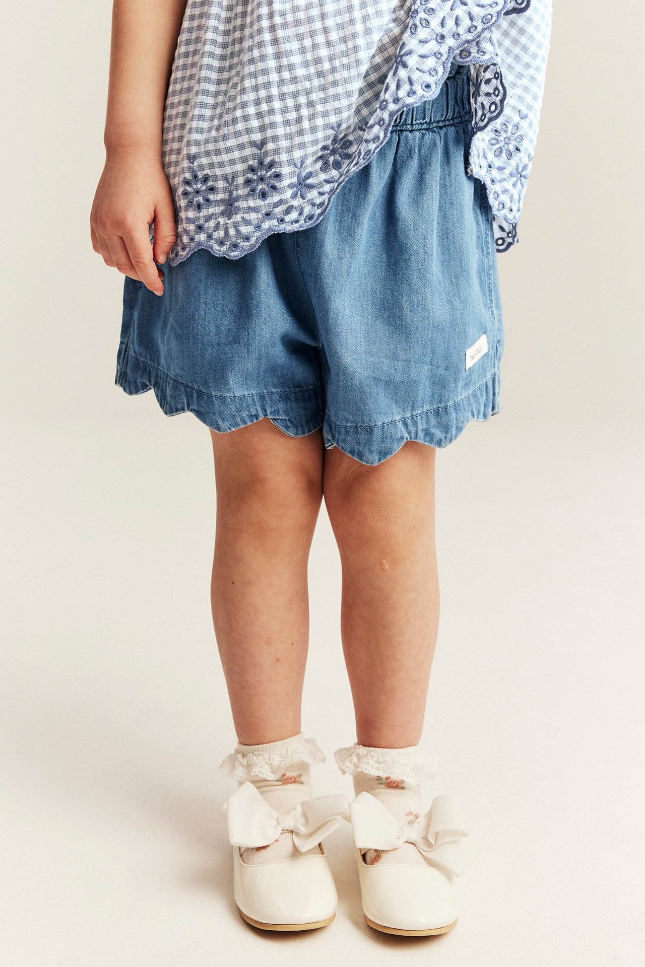 Front view of a kid wearing light denim shorts with a wavy edge, a blue checkered top, white lace socks, and white bow shoes.