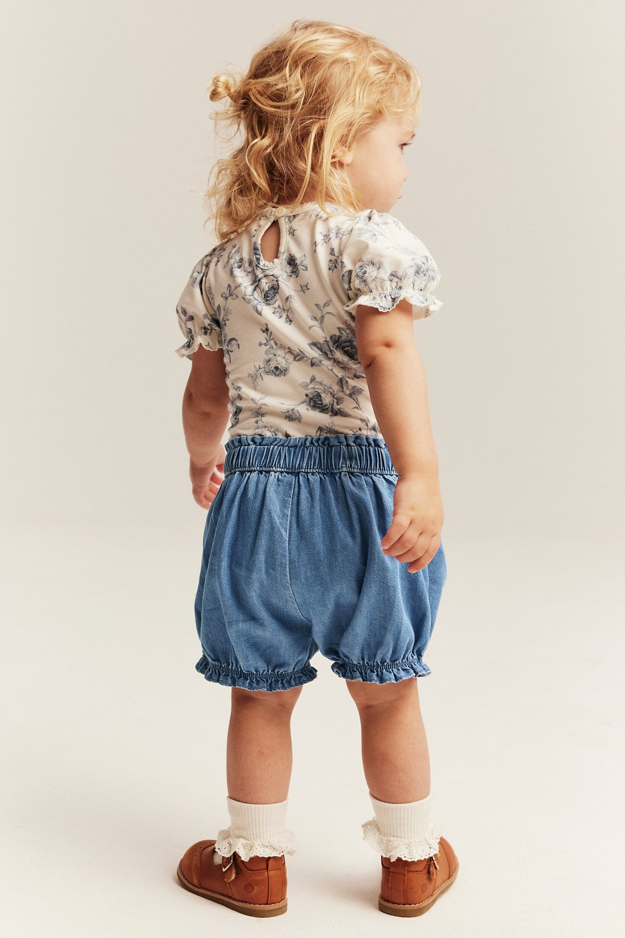 Rückenansicht eines Kindes in hellblauen Jeansshorts mit Rüschen, einem weißen Blümchen-Oberteil und braunen Schuhen.