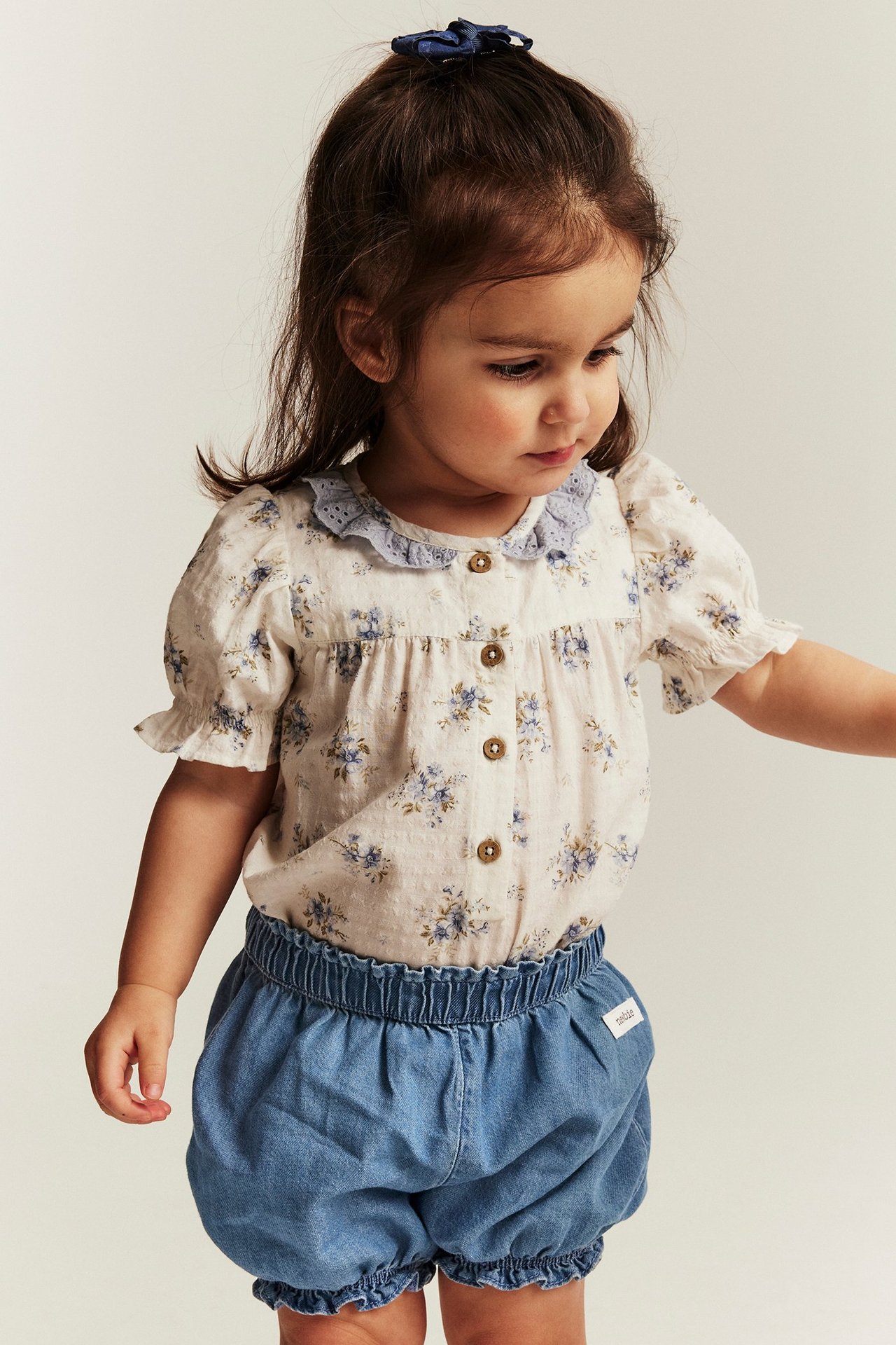 Vorderansicht: Kind trägt hellblaue Jeansshorts mit Rüschen, geblümte Bluse und blaue Haarschleife.