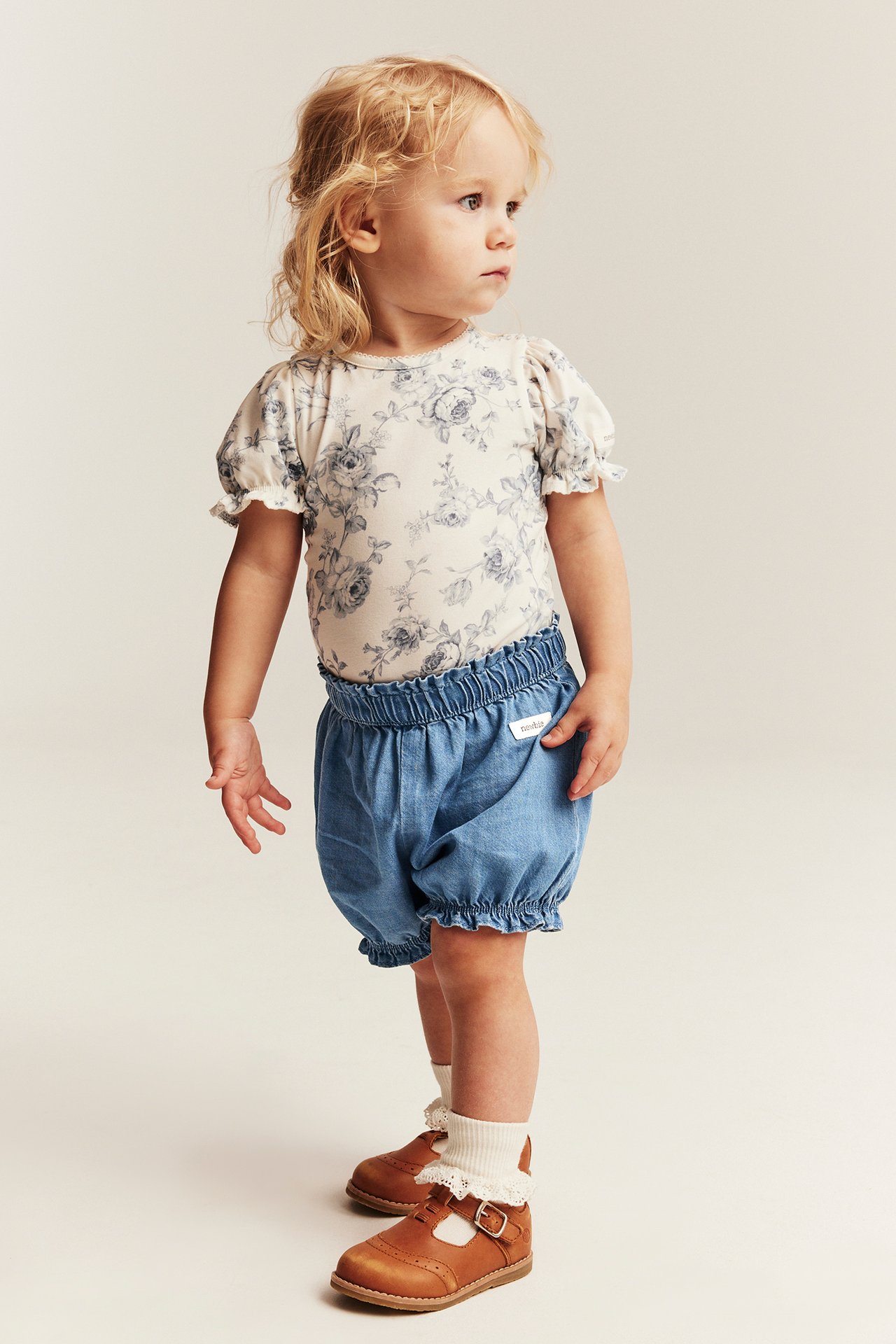Vorderansicht: Baby trägt hellblaue gewebte Jeans-Shorts, ein weißes Blumentop, braune Schuhe und weiße Rüschenstrümpfe.