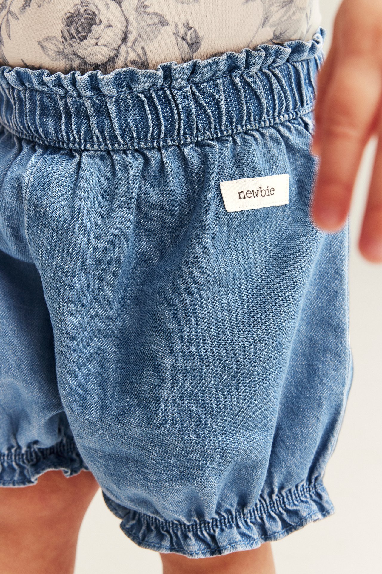 Nahaufnahme, Vorderansicht: Baby trägt hellblaue Jeans-Shorts mit gerafftem Gummibund an Taille und Beinenden, über einem Blumentop.