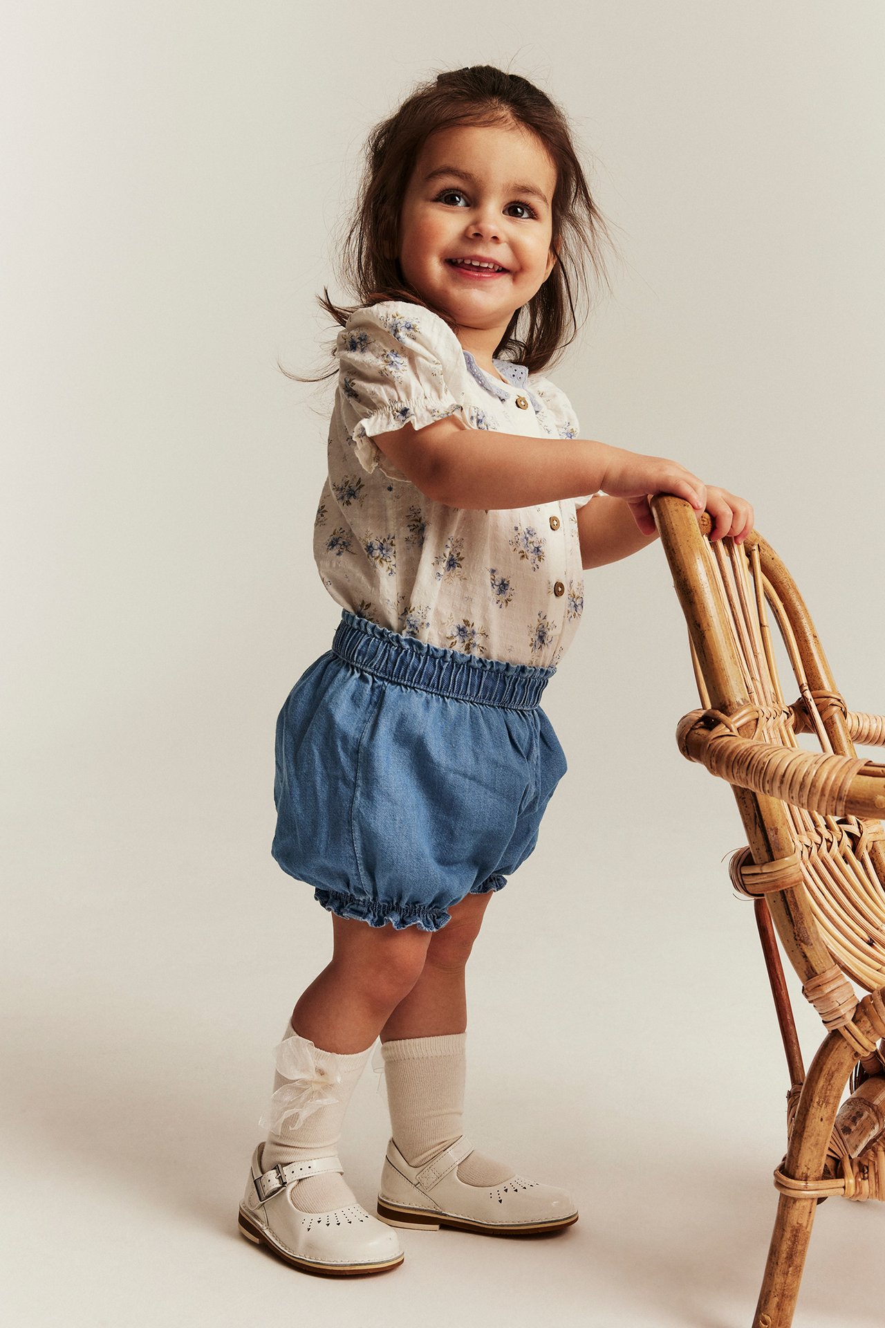 Vorderansicht: Lächelndes Kind in hellblauen Denim-Shorts mit Gummizug, geblümter Bluse, weißen Socken und Schuhen.