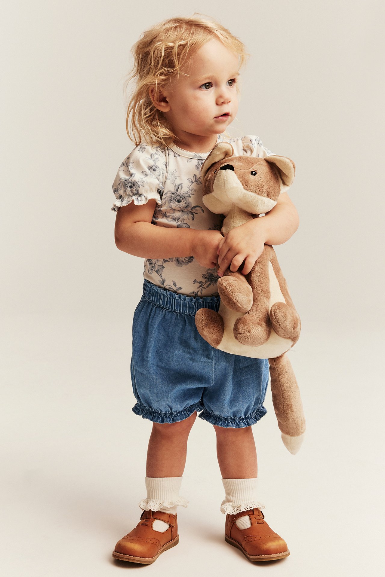 Front view: Baby girl in light blue denim shorts, ruffled hems, floral top, brown shoes, holding a toy.