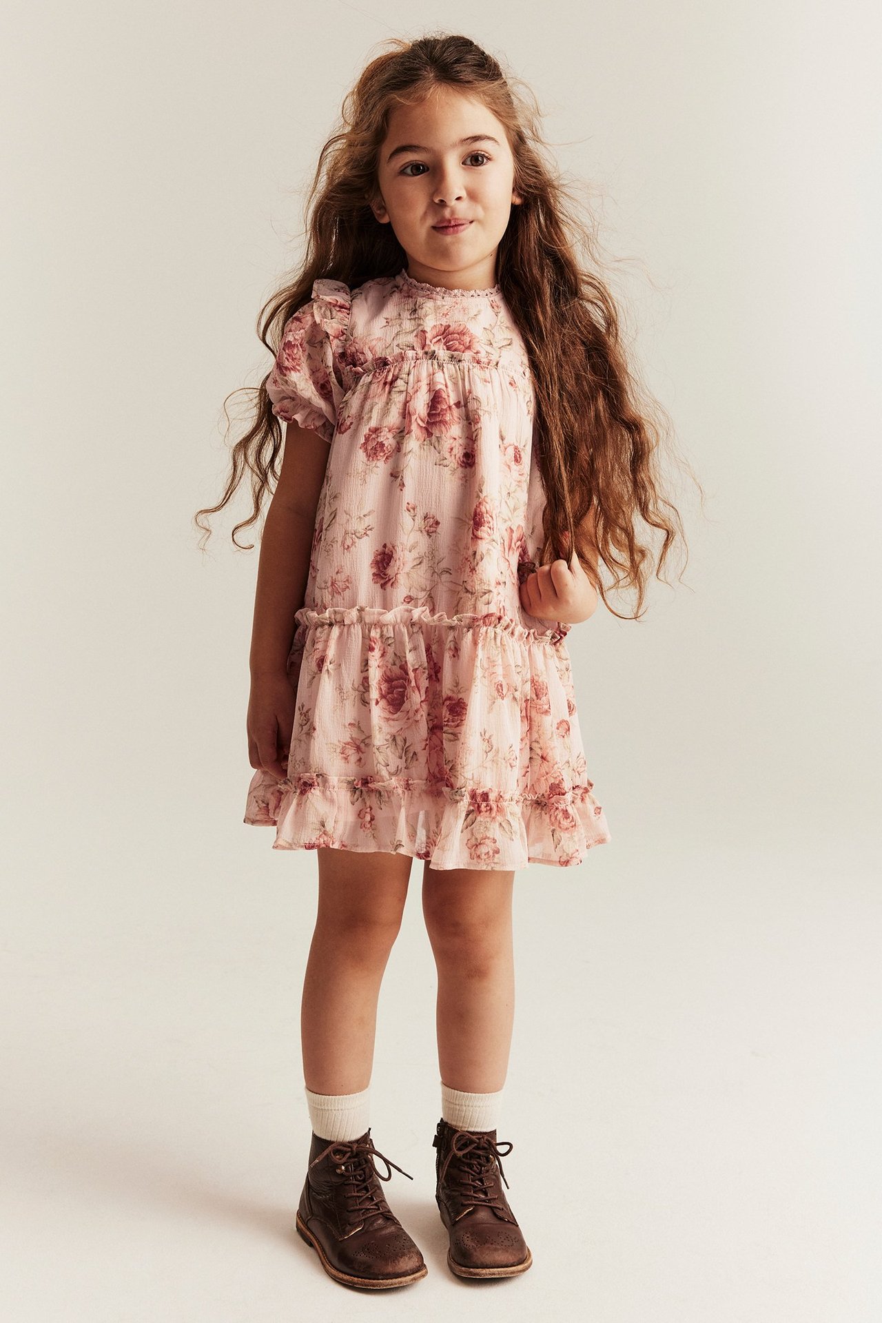 Front view of a kid wearing a pink floral chiffon dress with ruffles, white socks, and brown lace-up boots.