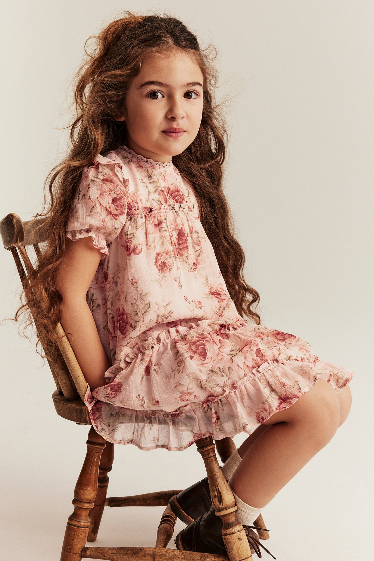 Front view of a kid sitting, wearing a pink floral chiffon dress with ruffles, white socks, and brown shoes.