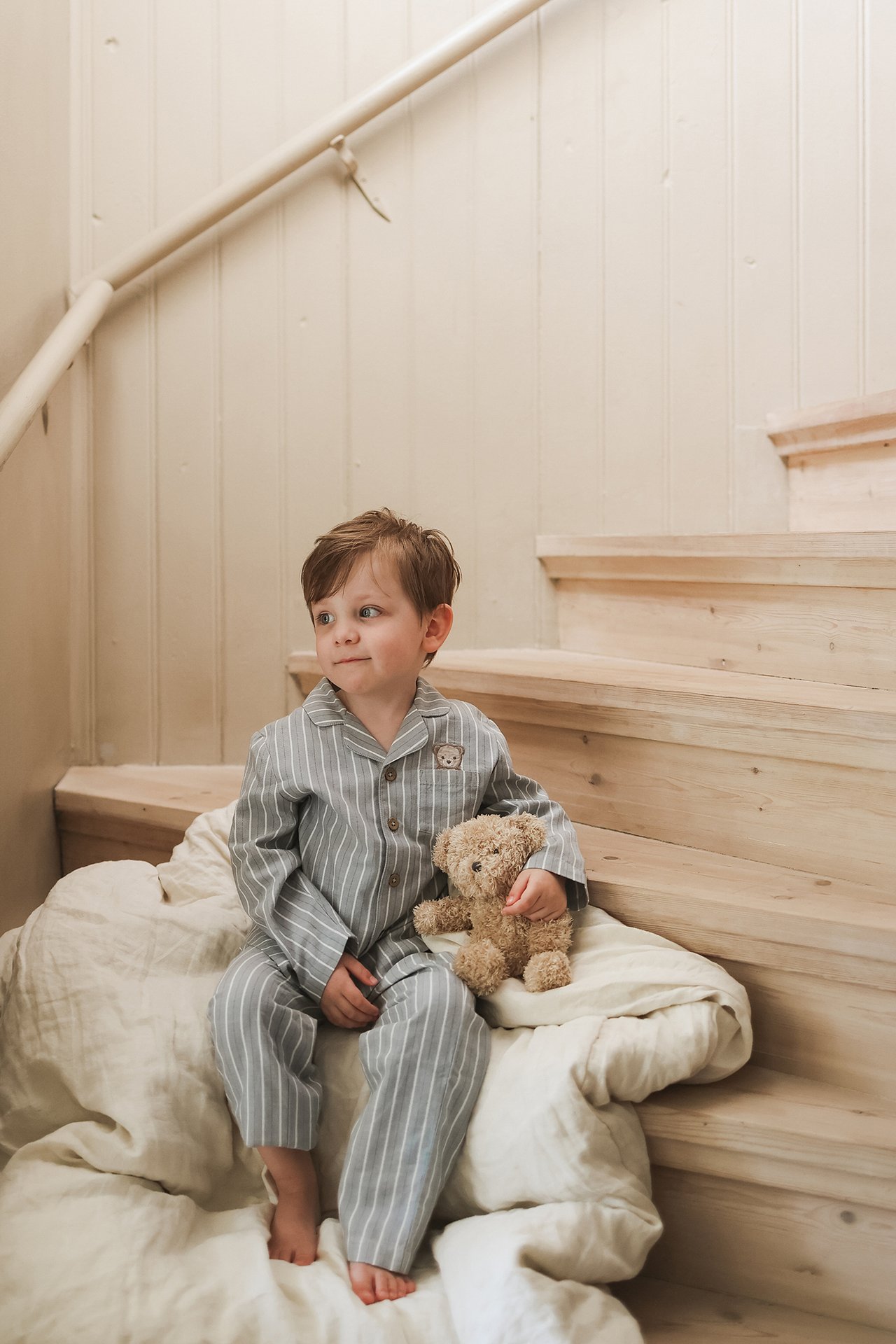 Seitenansicht eines Kindes in hellblau gestreiftem Baumwollpyjama mit Knöpfen, das einen Teddybären hält.
