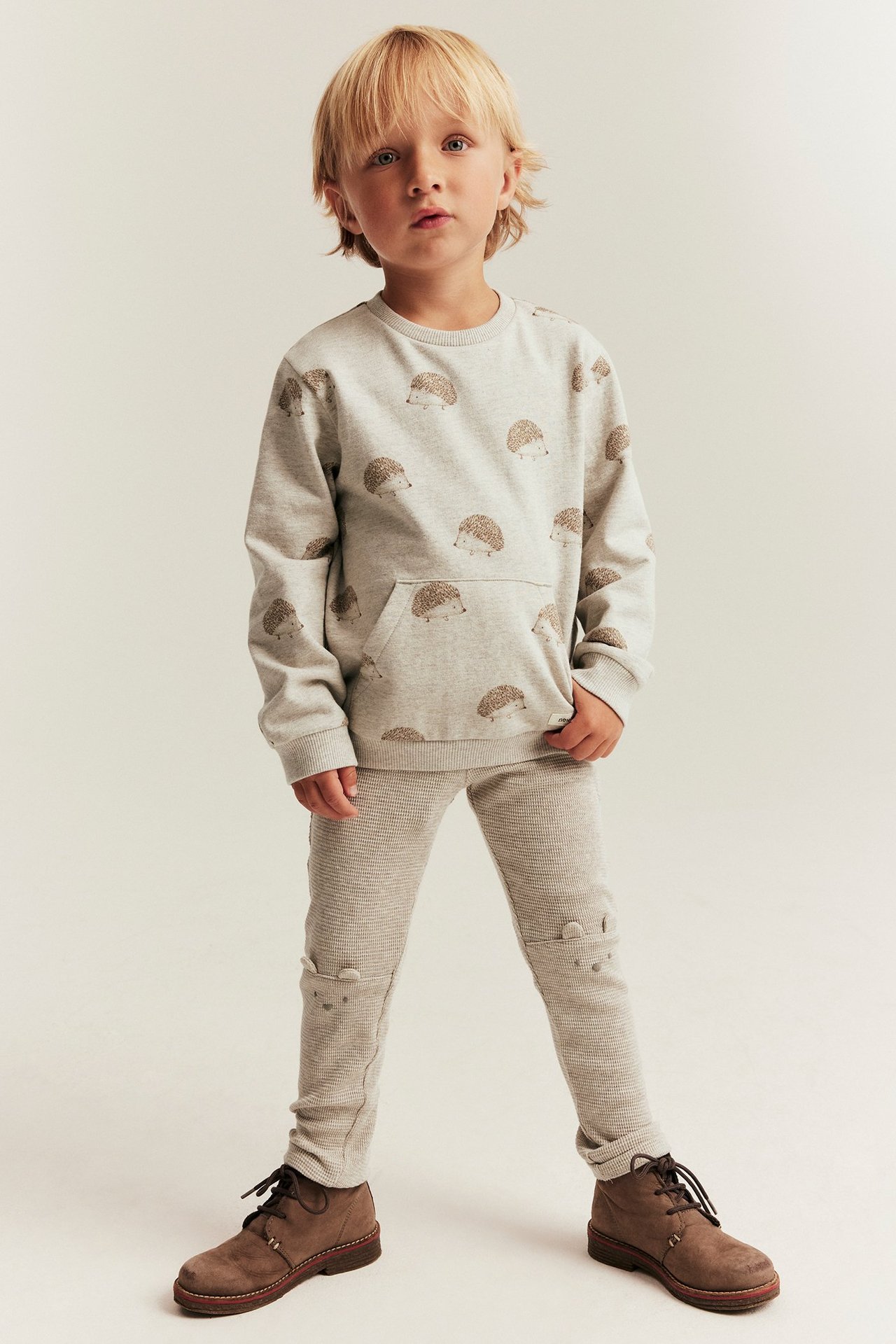Vorderansicht: Kind in hellbeigen Waffel-Leggings mit Knieohren, grauem Igel-Sweatshirt, braunen Stiefeln.