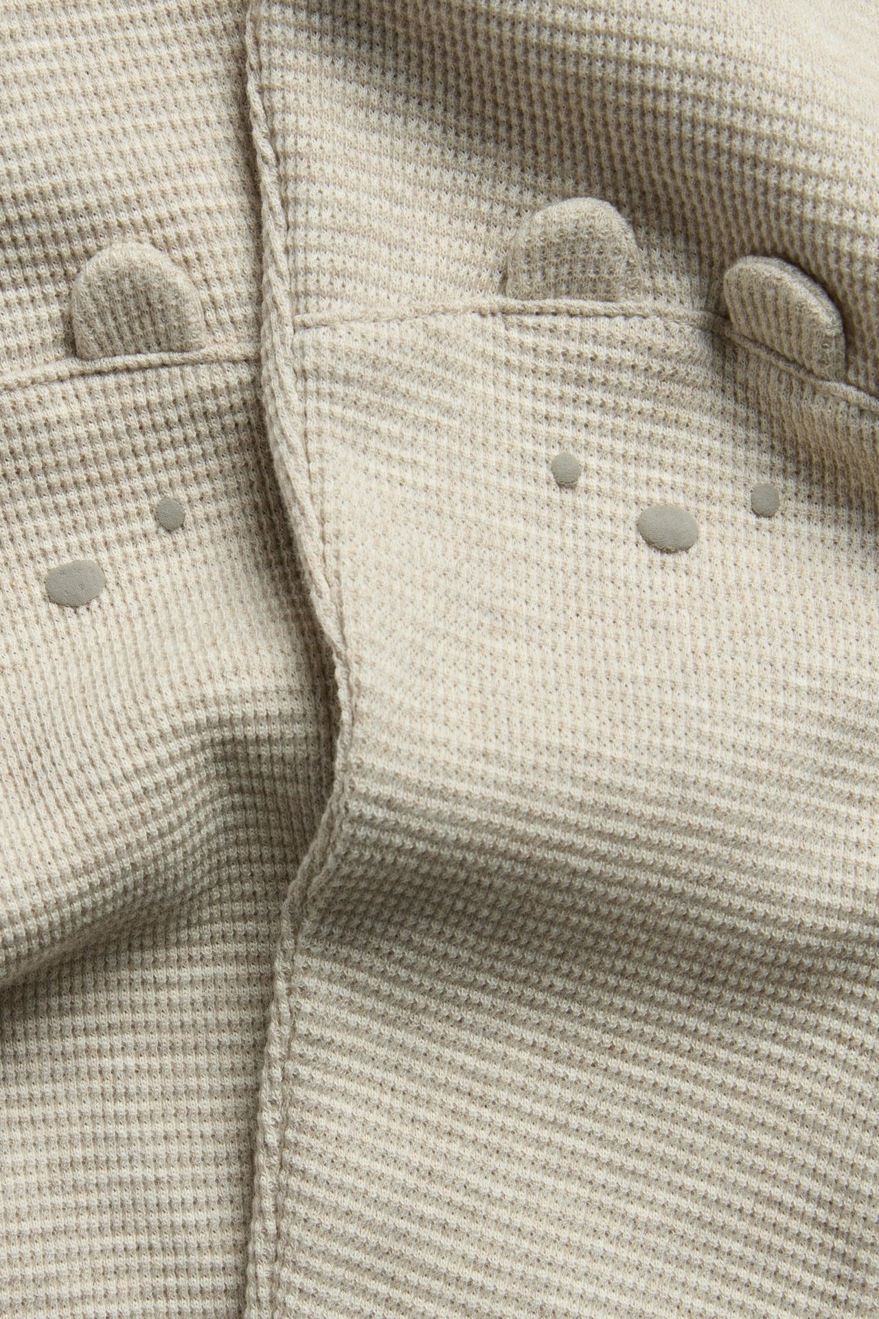 Nahaufnahme beigemelierter Waffelstrick-Leggings mit dekorativen Knöpfen vorne und kleinen Ohrenapplikationen.