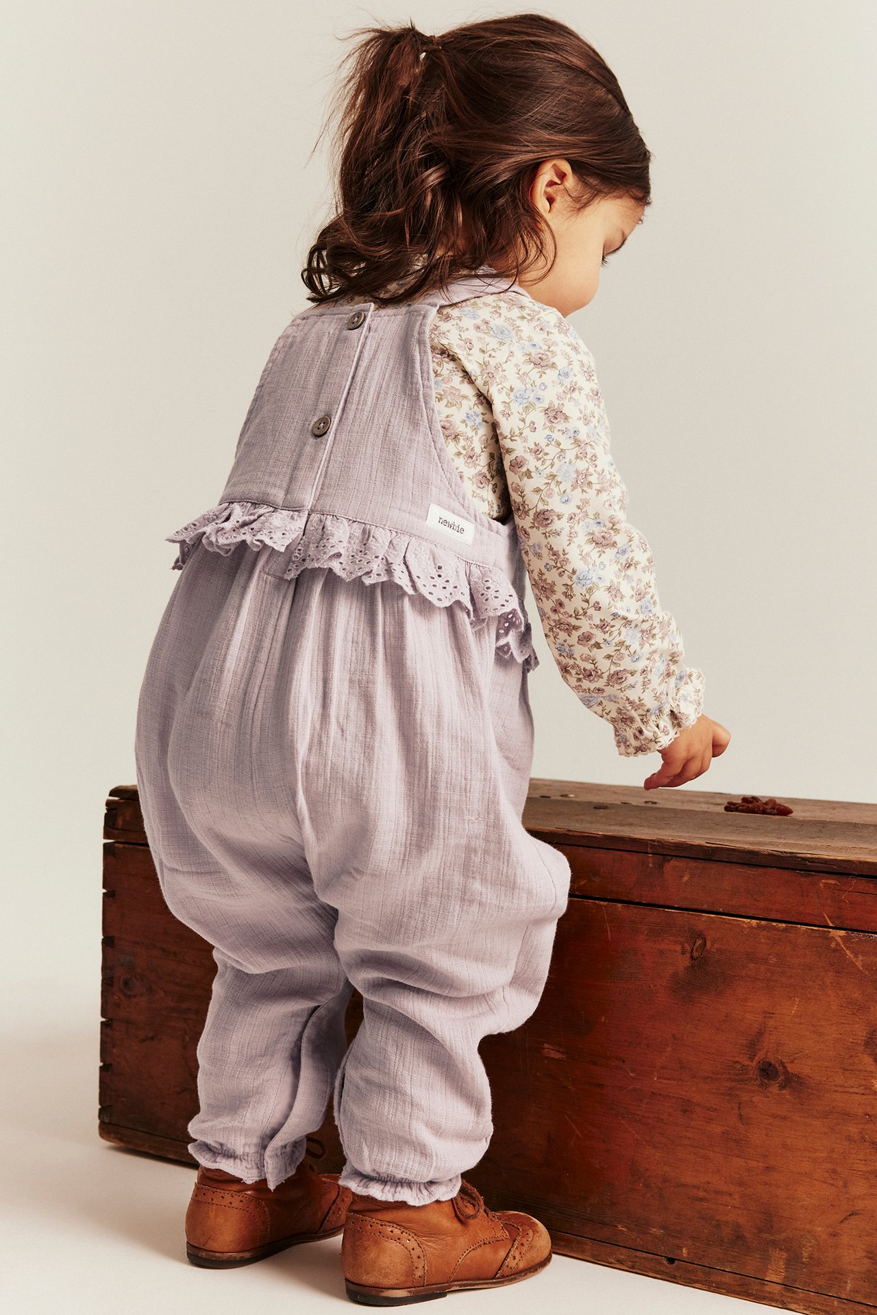 Back view: kid in light lilac cotton overalls with lace trim, floral shirt, and brown shoes.
