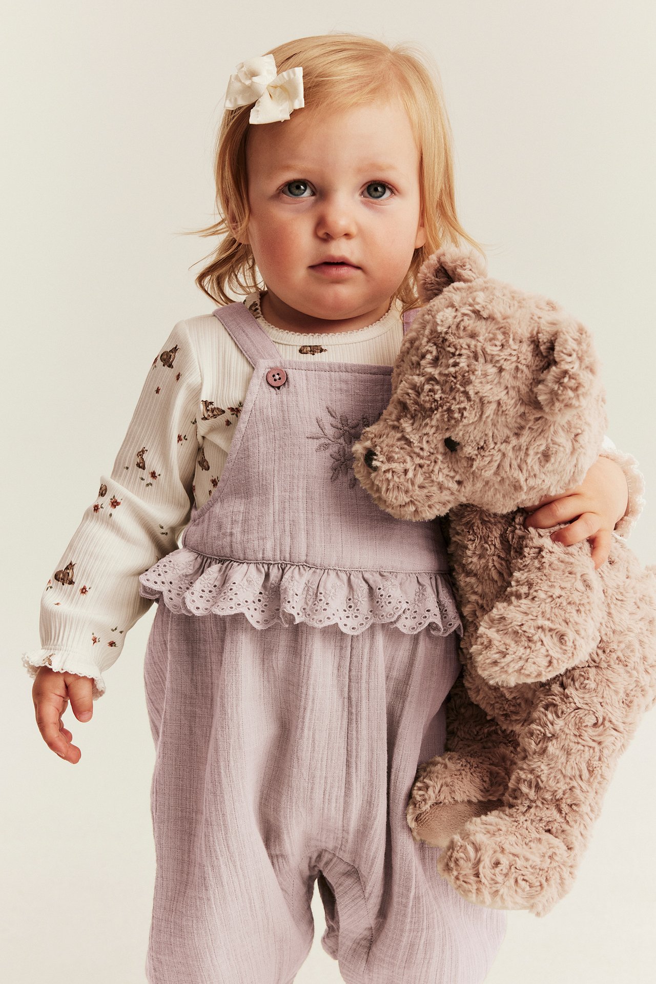 Baby in light lilac cotton overalls with lace ruffle and flower embroidery, holding a brown teddy bear.