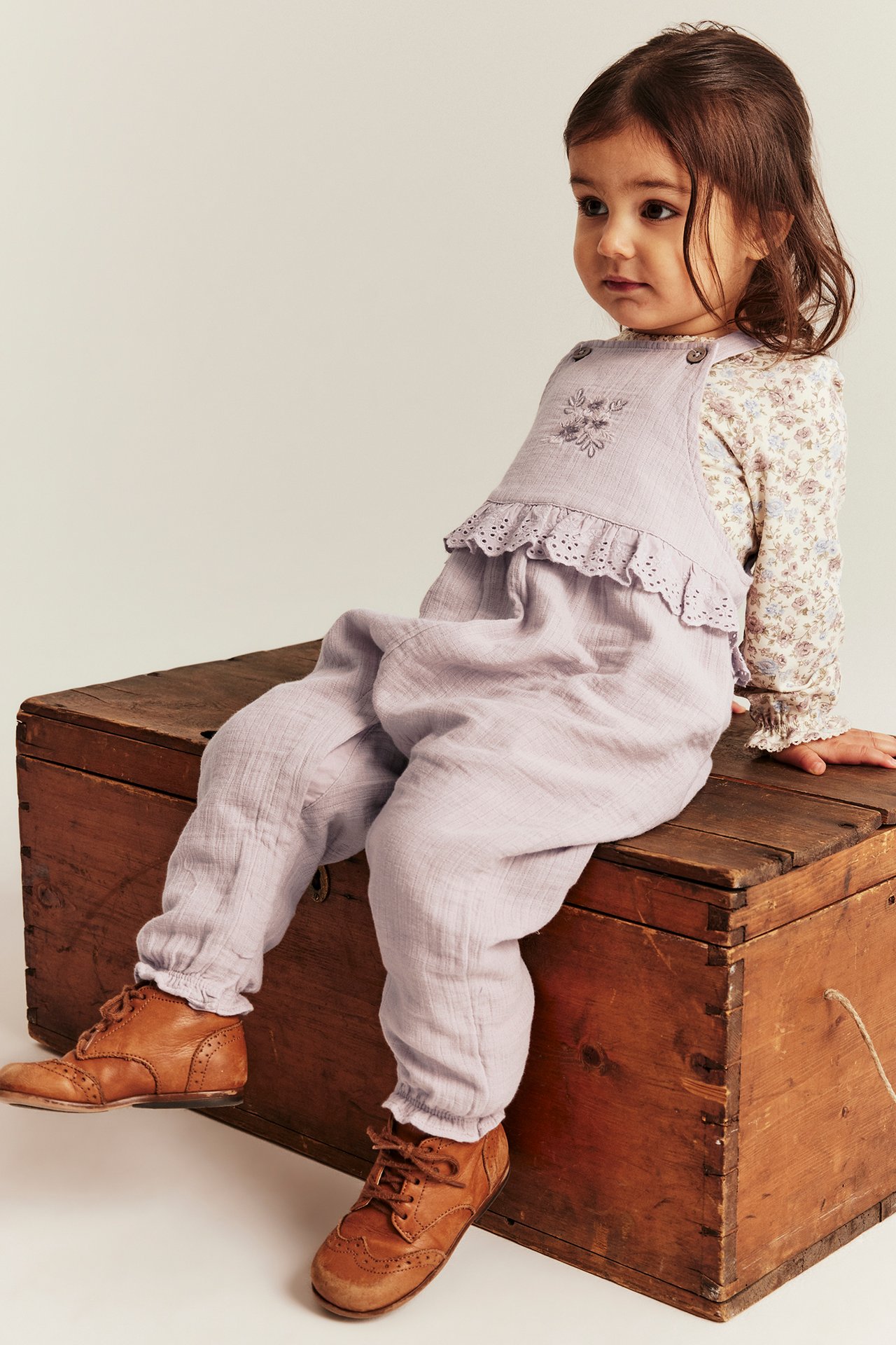 Front view: Kid in light lilac overalls with lace, flower embroidery, over a floral shirt, wearing brown shoes.