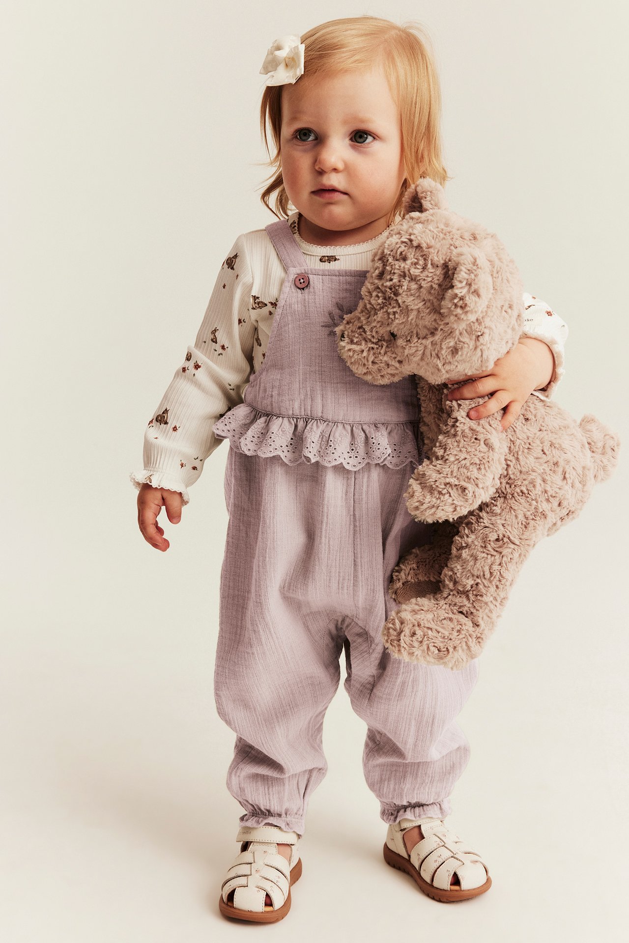 Vorderansicht: Baby trägt helllilafarbene Baumwoll-Latzhose mit Spitze, über gemustertem Oberteil, hält einen braunen Teddy.