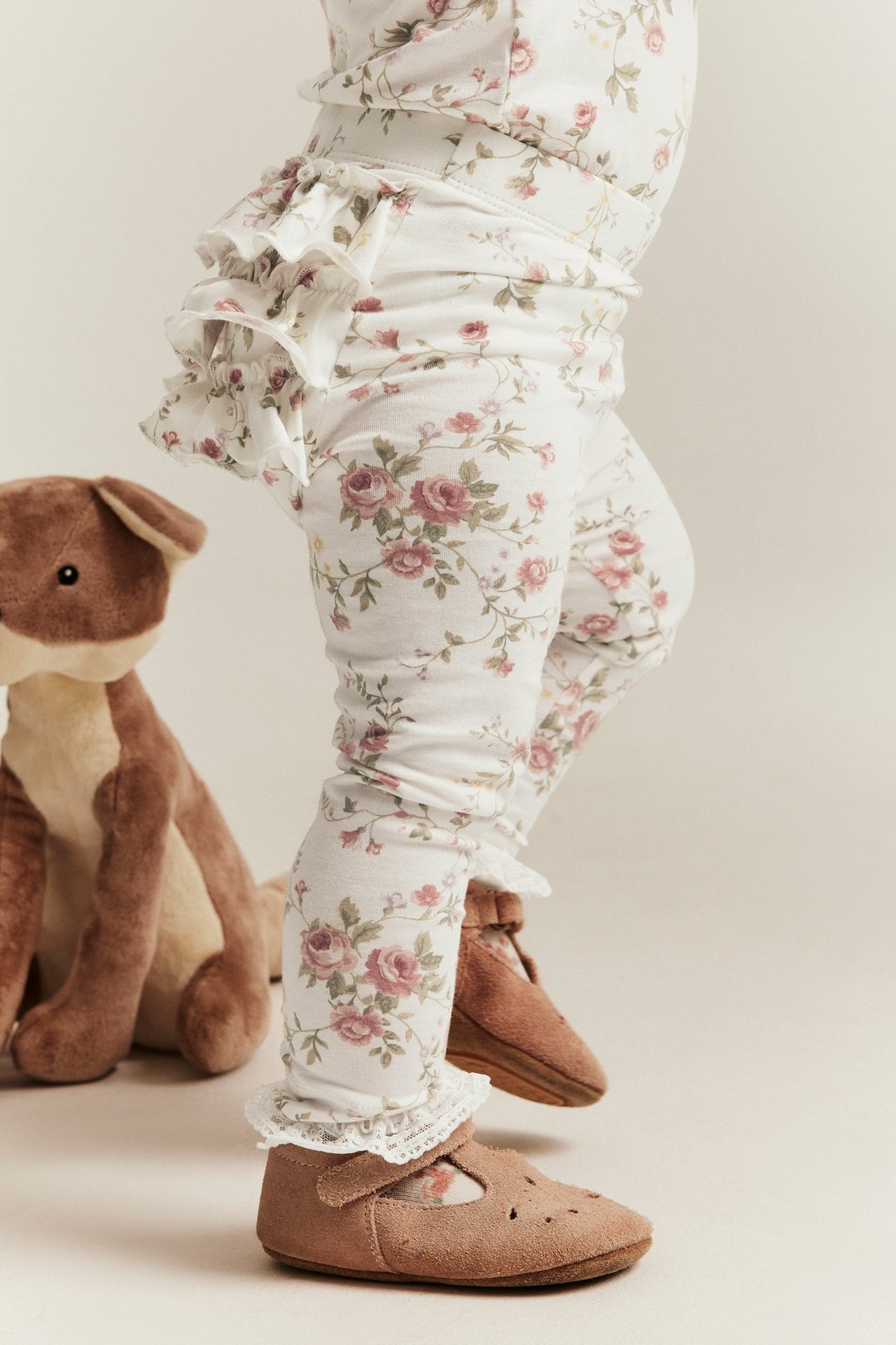 Side view of a baby wearing off-white floral leggings with back ruffles and brown suede shoes.