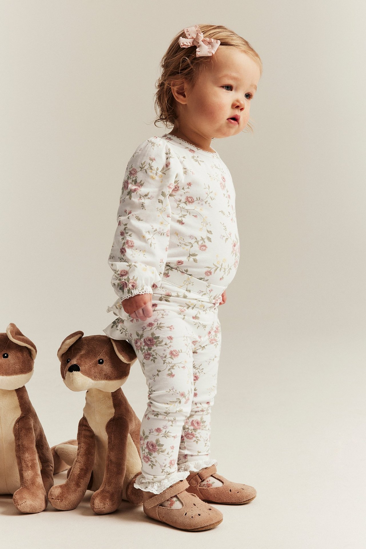 Side view of a baby wearing off-white floral leggings with ruffles, matching top, brown shoes, and a pink hair clip.
