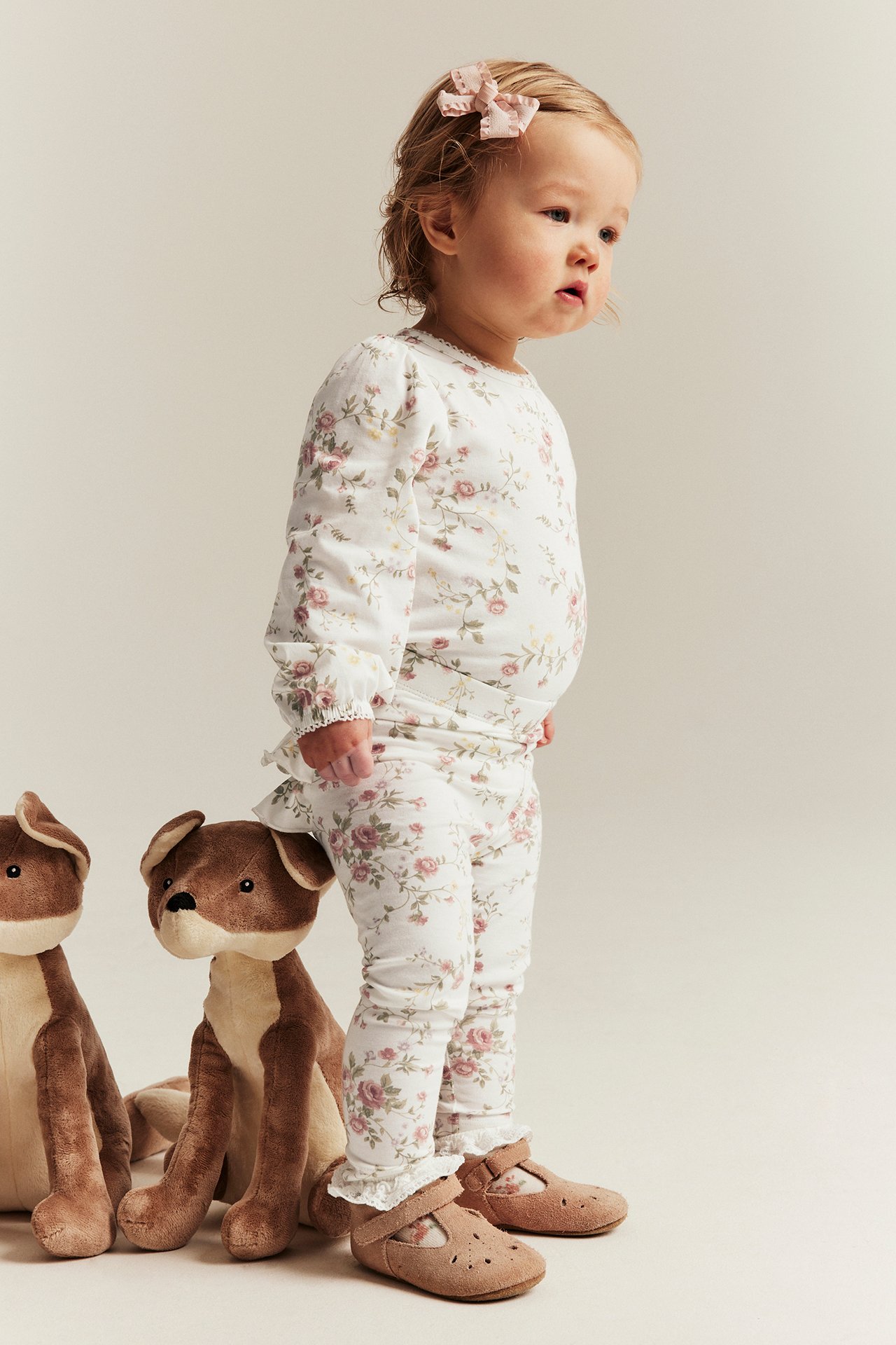 Side view of a baby wearing off-white floral leggings with ruffles, matching top, brown shoes, and a pink hair clip.