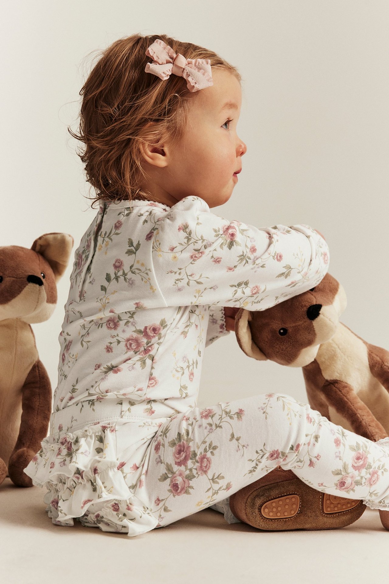 Side view of baby in off-white floral ruffled leggings, matching top, pink bow, and brown shoes.