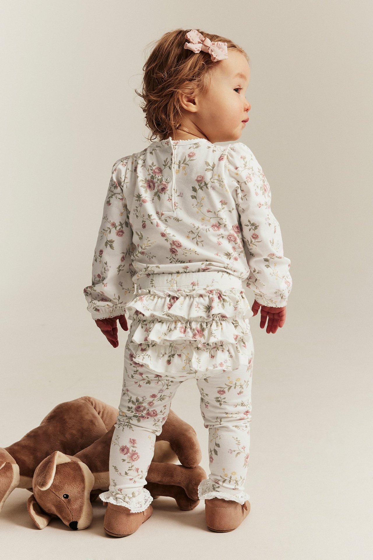 Rückenansicht eines Babys in cremefarbenen Blumen-Leggings mit Rüschen, passendem Oberteil, braunen Schuhen und rosa Schleife.