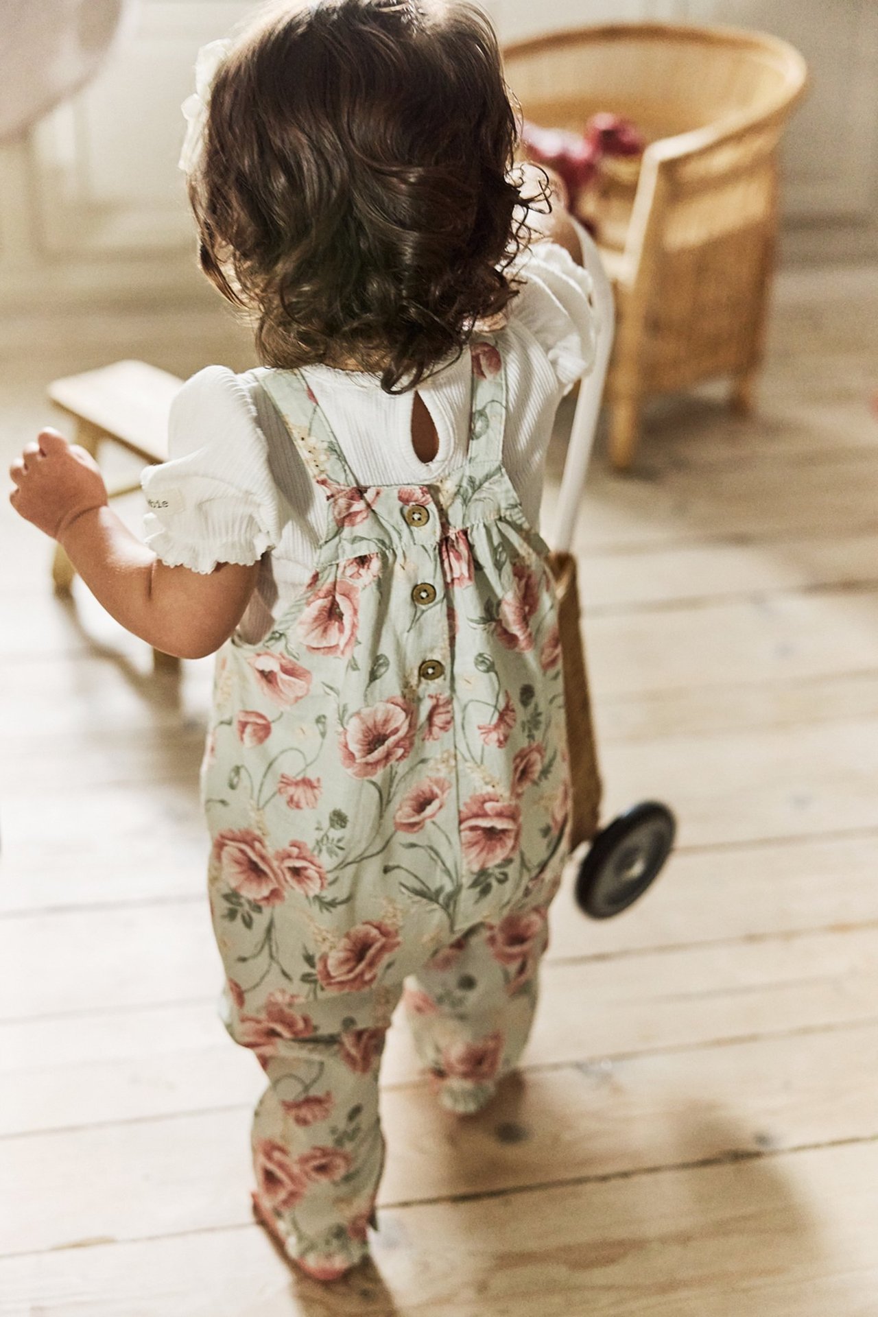 Baby's back view: light green floral cotton dungarees with back buttons over a white ribbed top.