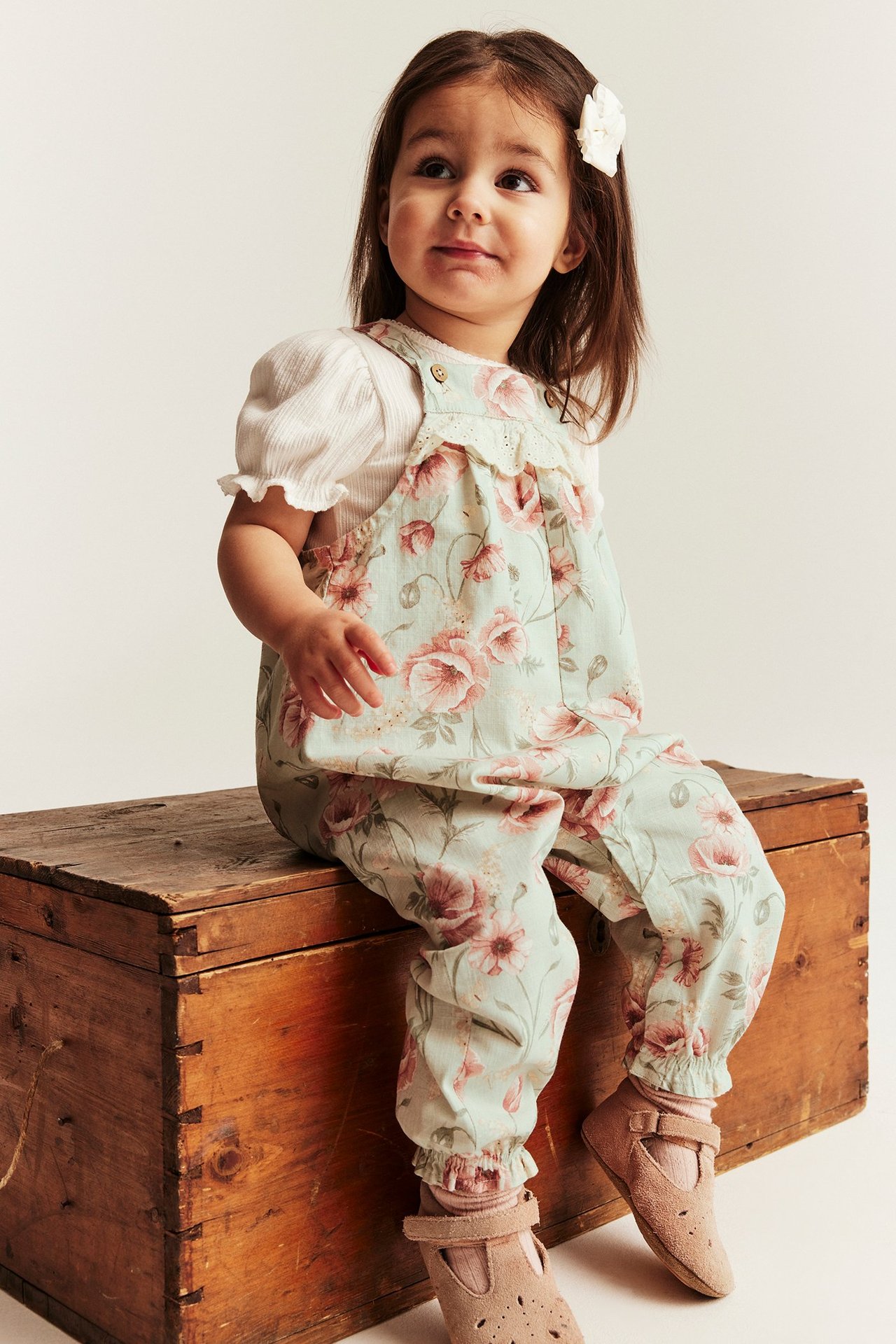 Front view: Kid in light green floral cotton dungarees, white top, pink socks, and brown shoes.