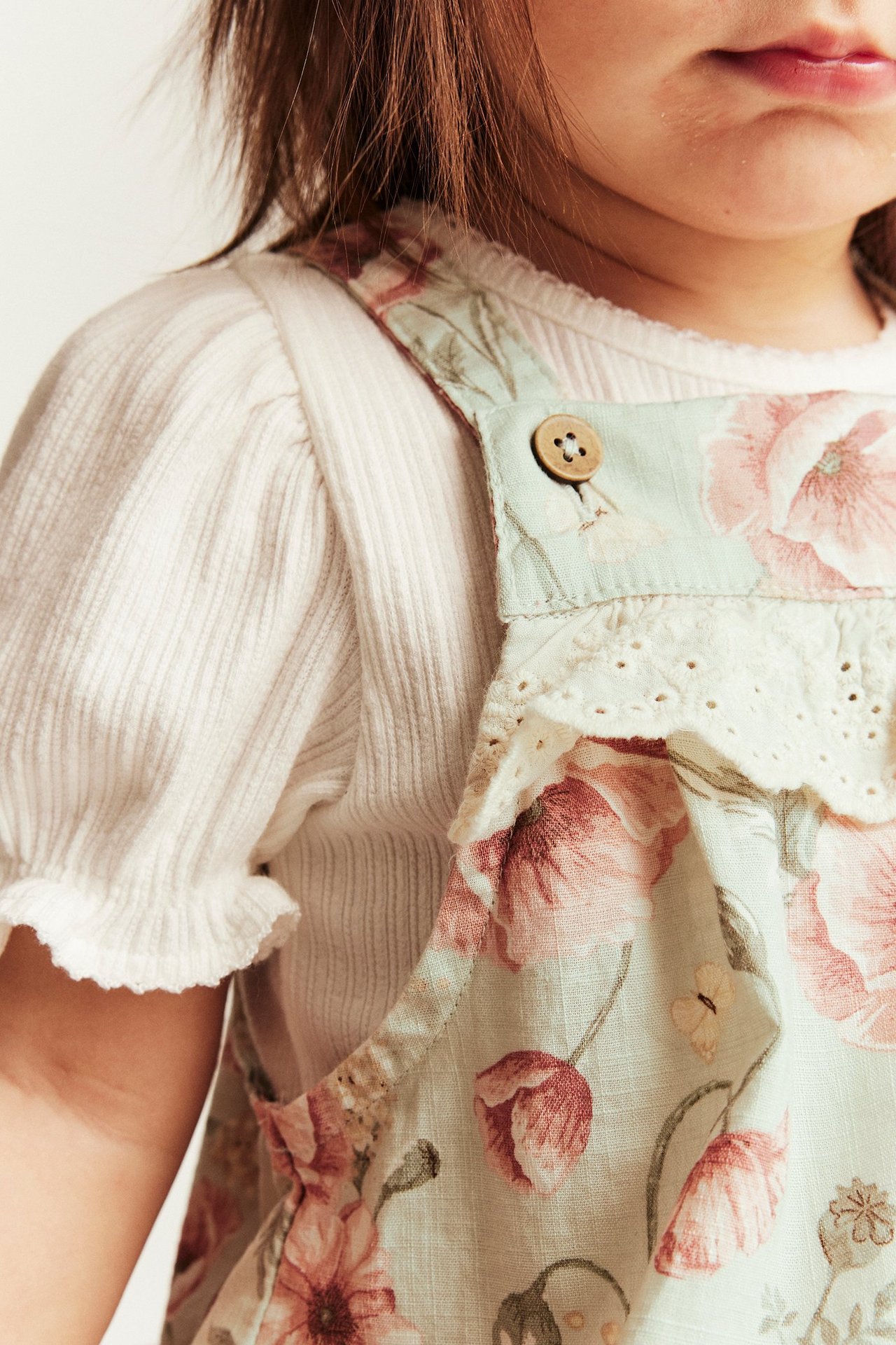 Close-up of a kid wearing light green floral dungarees with a lace ruffle over a white ribbed top.