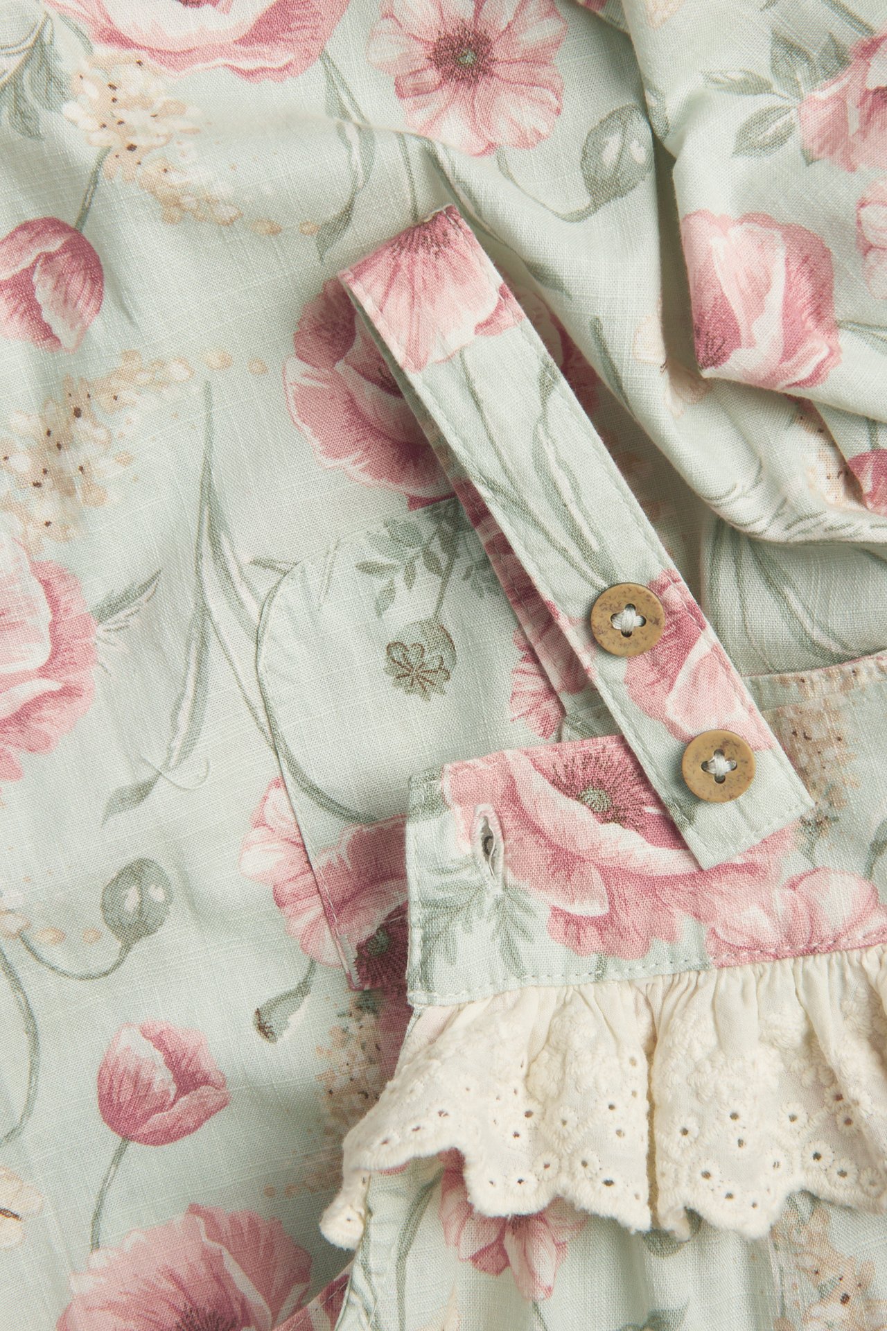 Close-up of kid's light green cotton floral dungarees with pink flowers, lace ruffle, and wooden buttons.