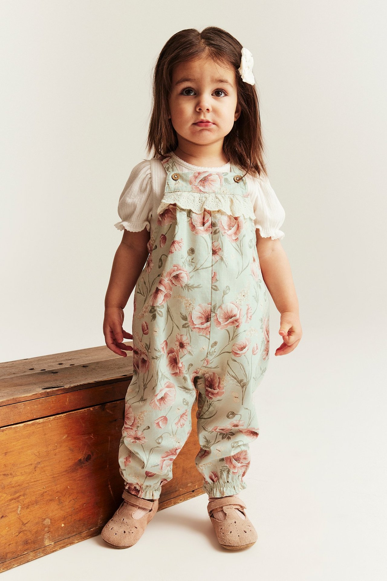 Front view of a kid in light green floral cotton dungarees over a white ribbed top, with brown shoes.