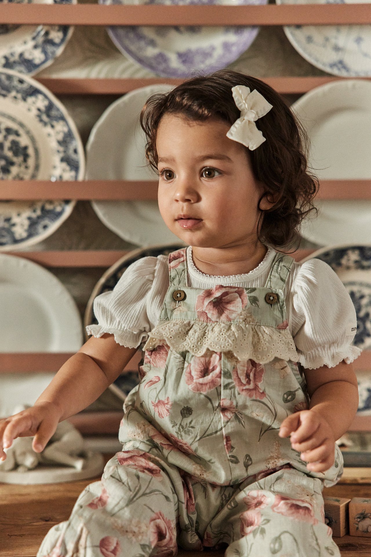 Front view of a baby wearing light green floral dungarees with lace ruffle, a white textured top, and a white bow clip.