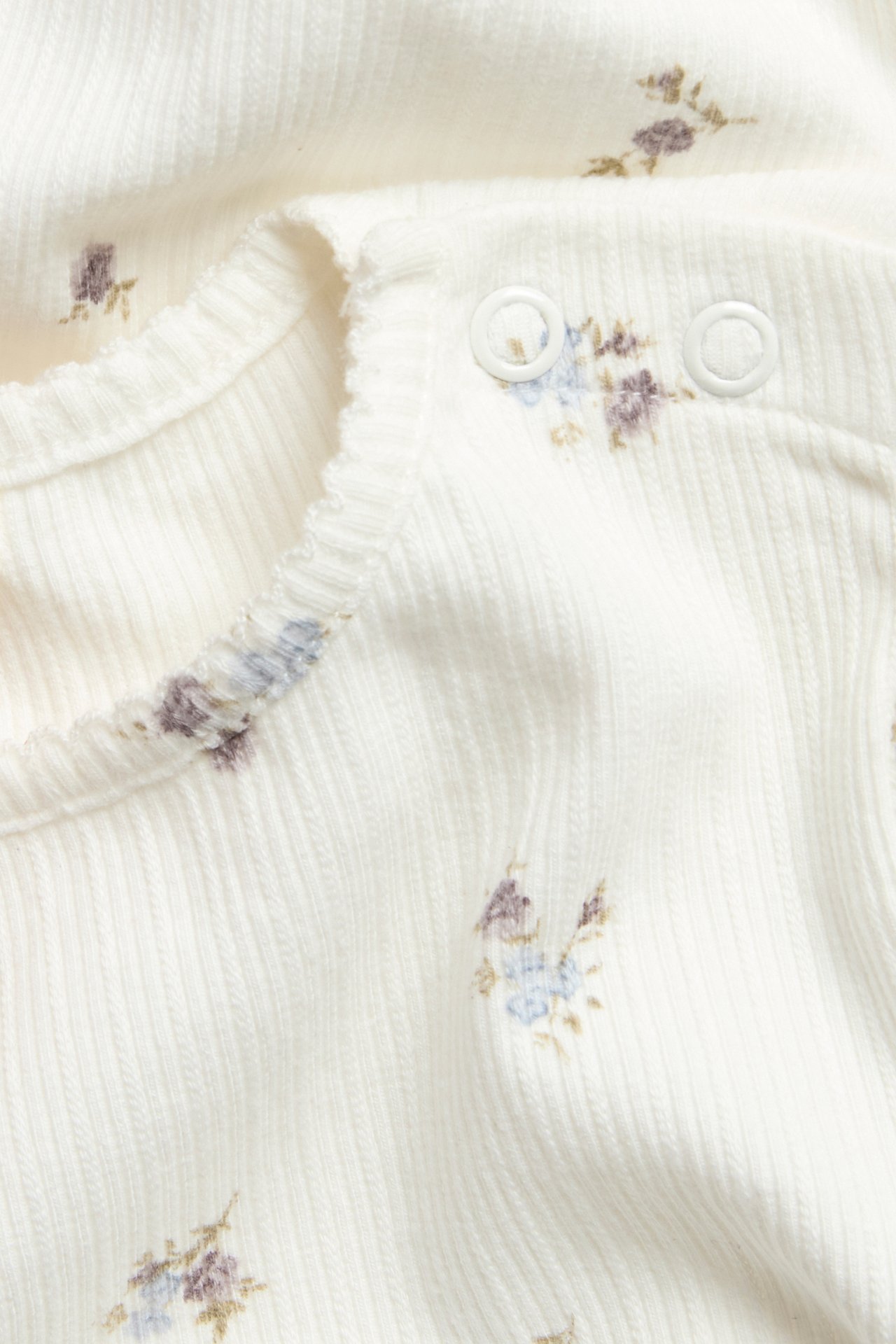 Close-up of an off-white ribbed baby bodysuit with a floral print, picot trim, and snap buttons.
