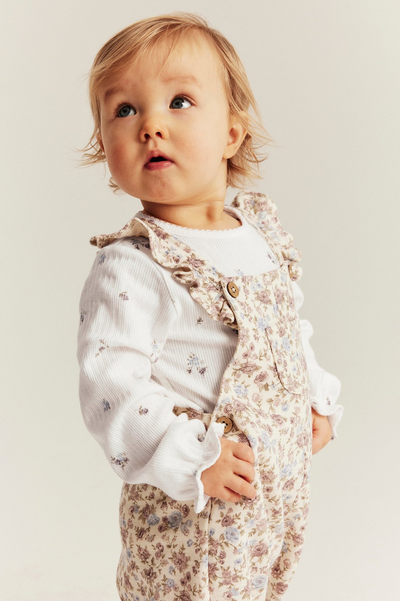 Vorderansicht eines Babys in einem cremeweißen floralen Ripp-Body mit Rüschenbündchen unter einem floralen Overall.