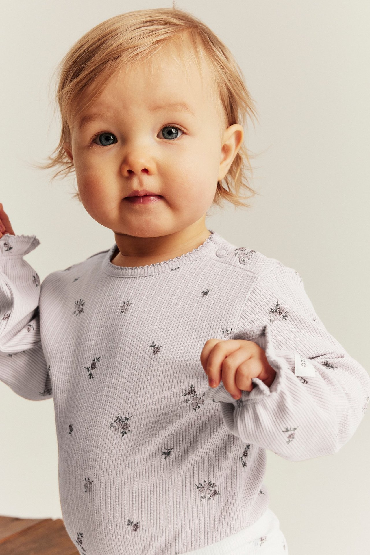 Nahaufnahme von vorne eines Babys in helllila geripptem Body mit Blumenmuster, Rüschen und Druckknöpfen.