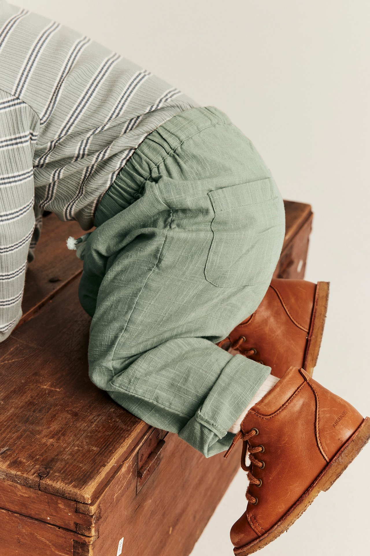 Close-up back view of baby in dark green structured cotton trousers, striped top, and brown boots.