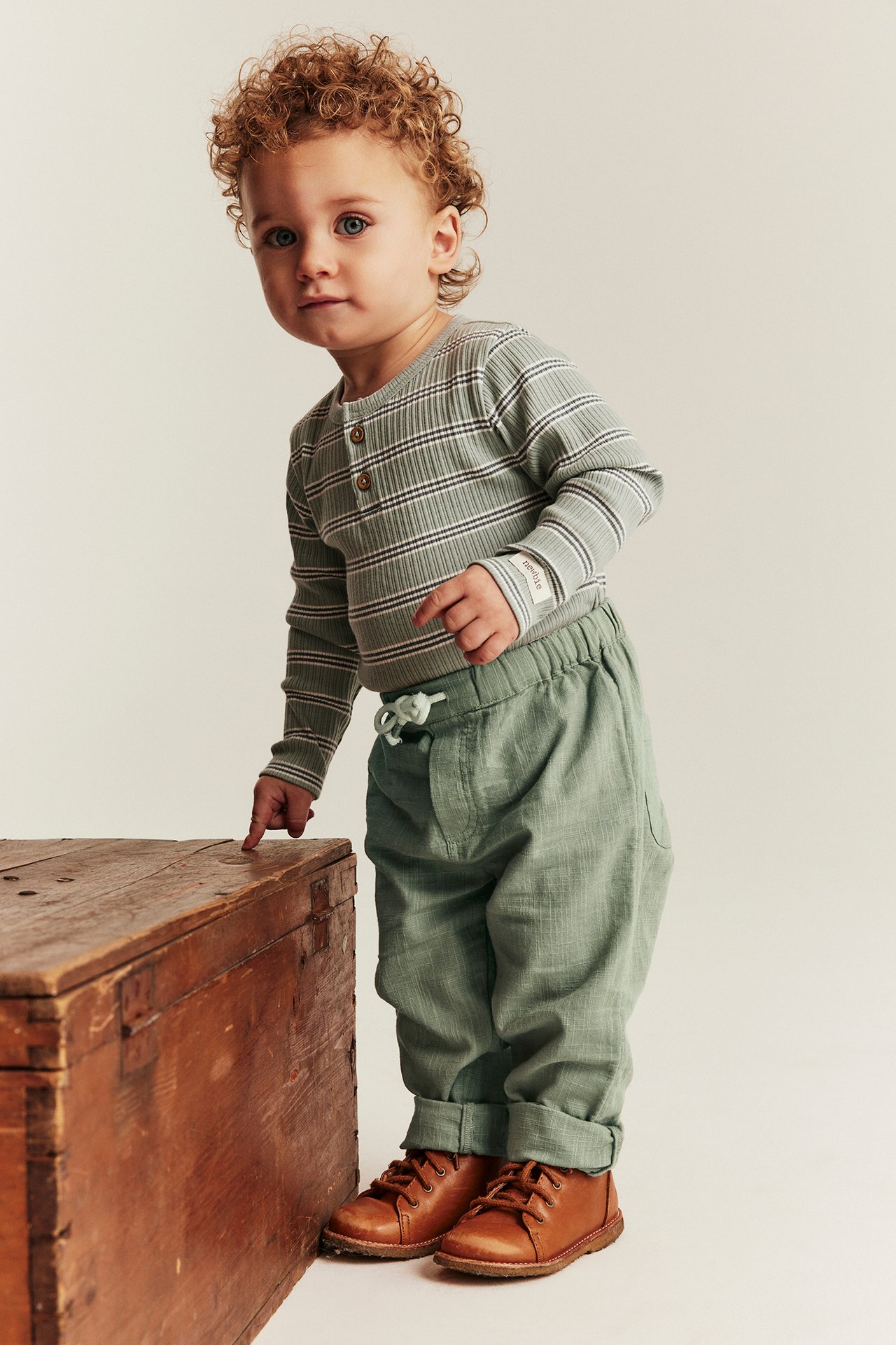 Front view: Baby wearing dark green textured woven trousers with cuffed hems and drawstring, a striped bodysuit, and brown boots.