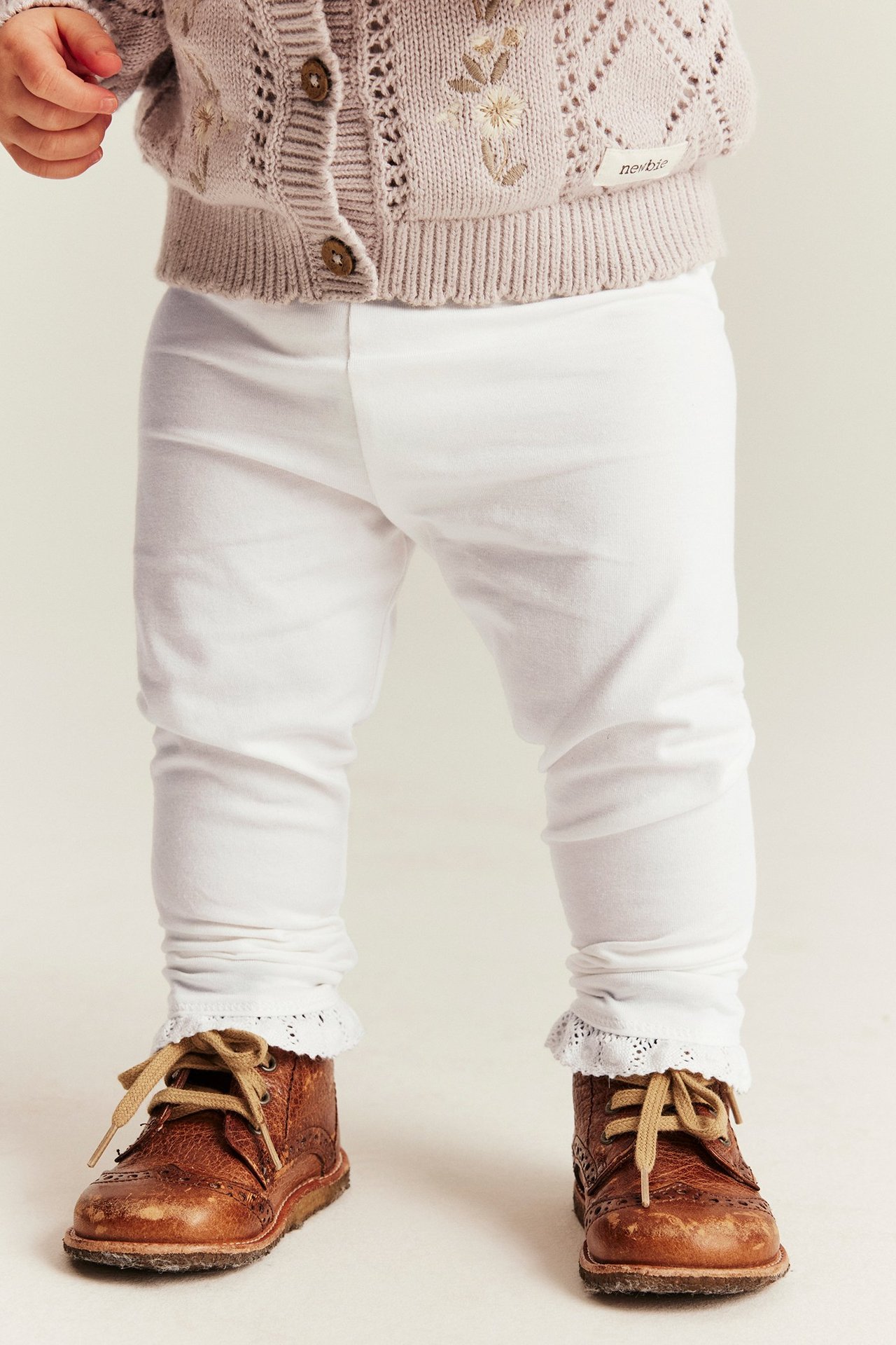 Front view of baby wearing offwhite stretchy leggings with lace trim, light beige knitted cardigan, and brown boots.