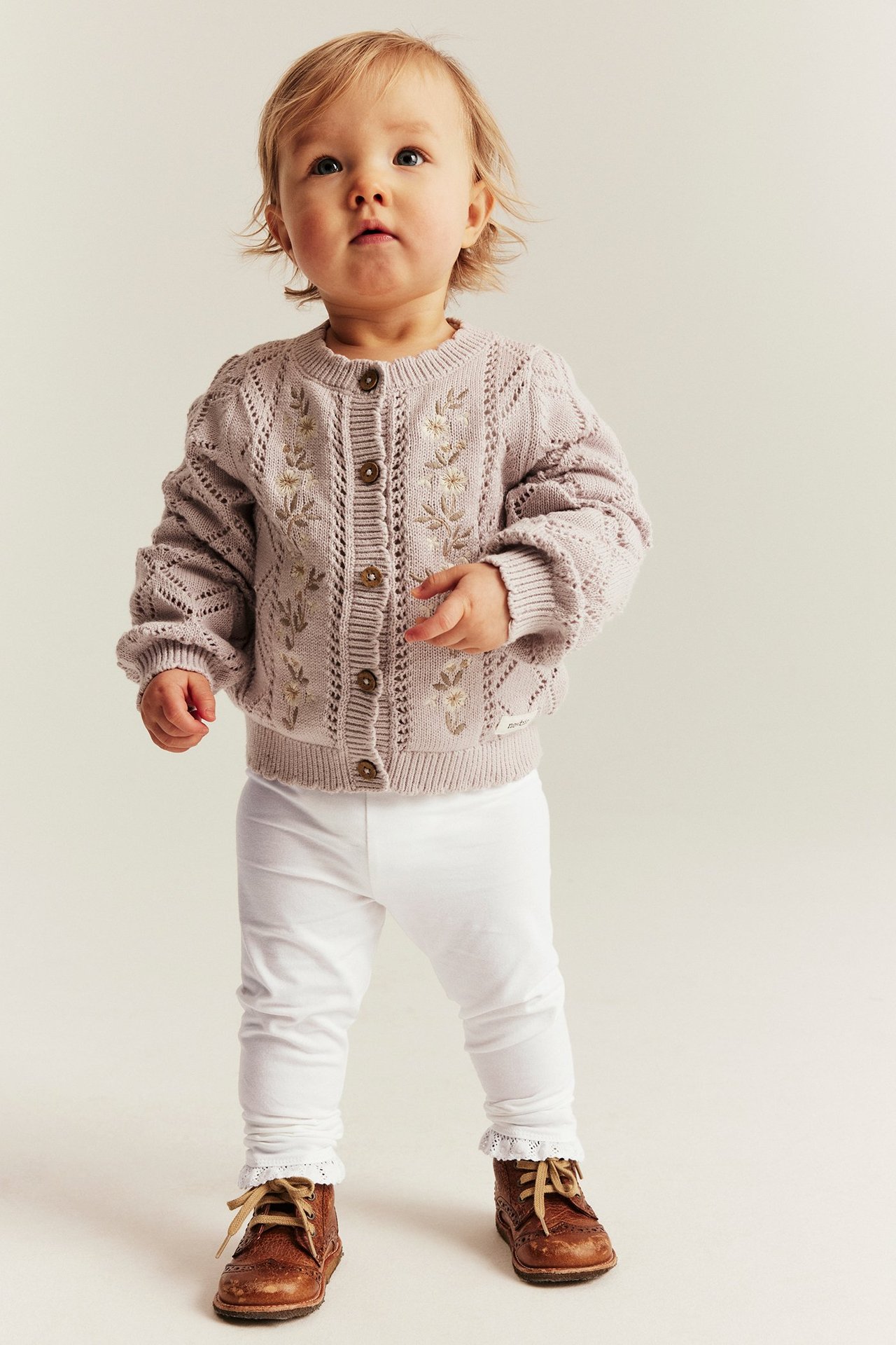 Vorderansicht eines Babys in cremeweißen Stretch-Leggings mit Spitzensaum, einem lila bestickten Cardigan und braunen Schuhen.