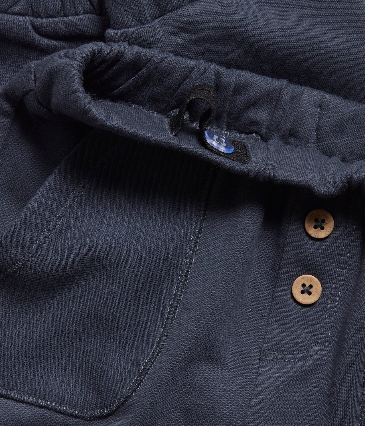 Close-up of dark blue cotton sweatpants with elastic waist, decorative wooden buttons, and ribbed patch pocket.