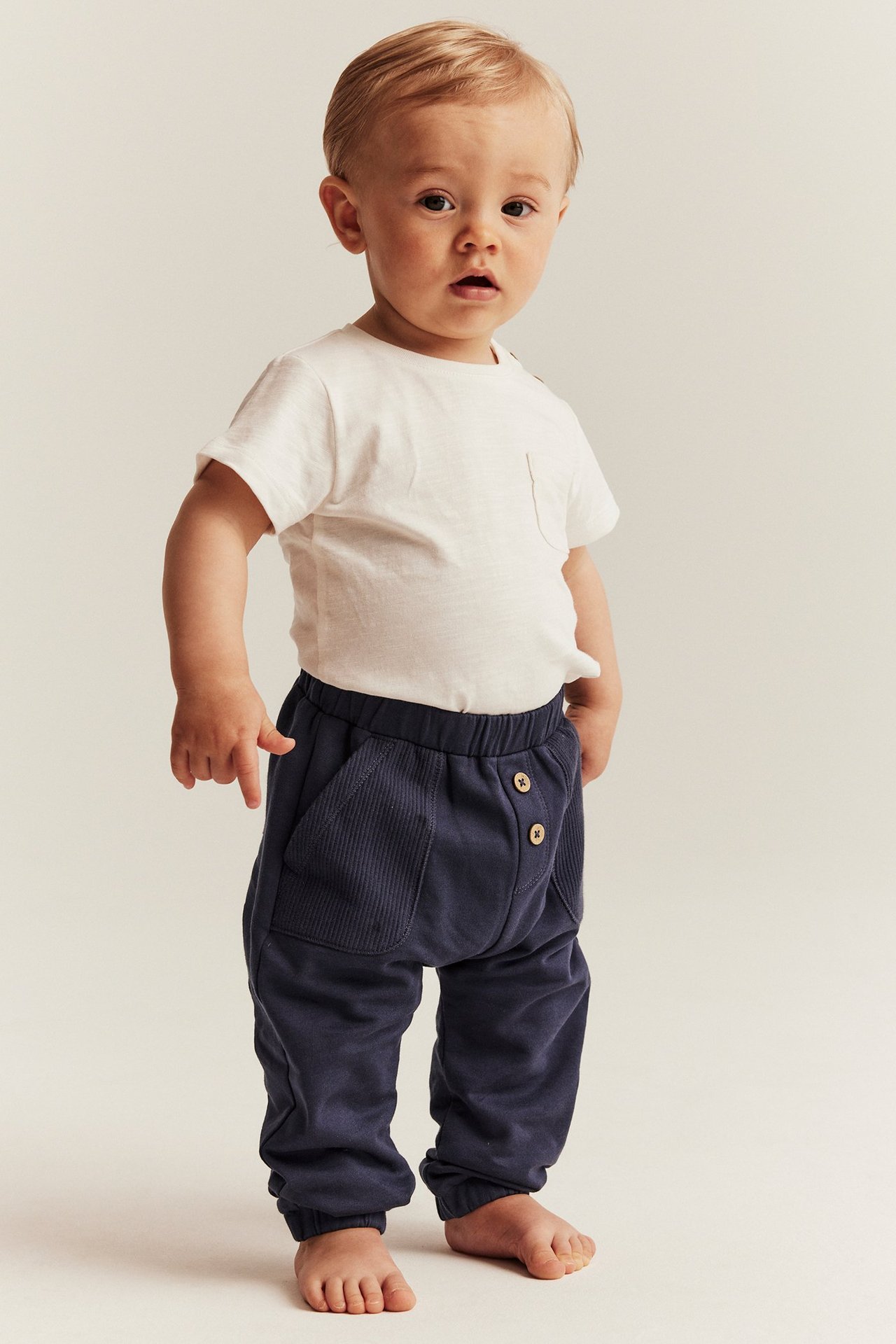 Front view of a baby wearing dark blue cotton jogging pants with ribbed pockets and a white t-shirt.