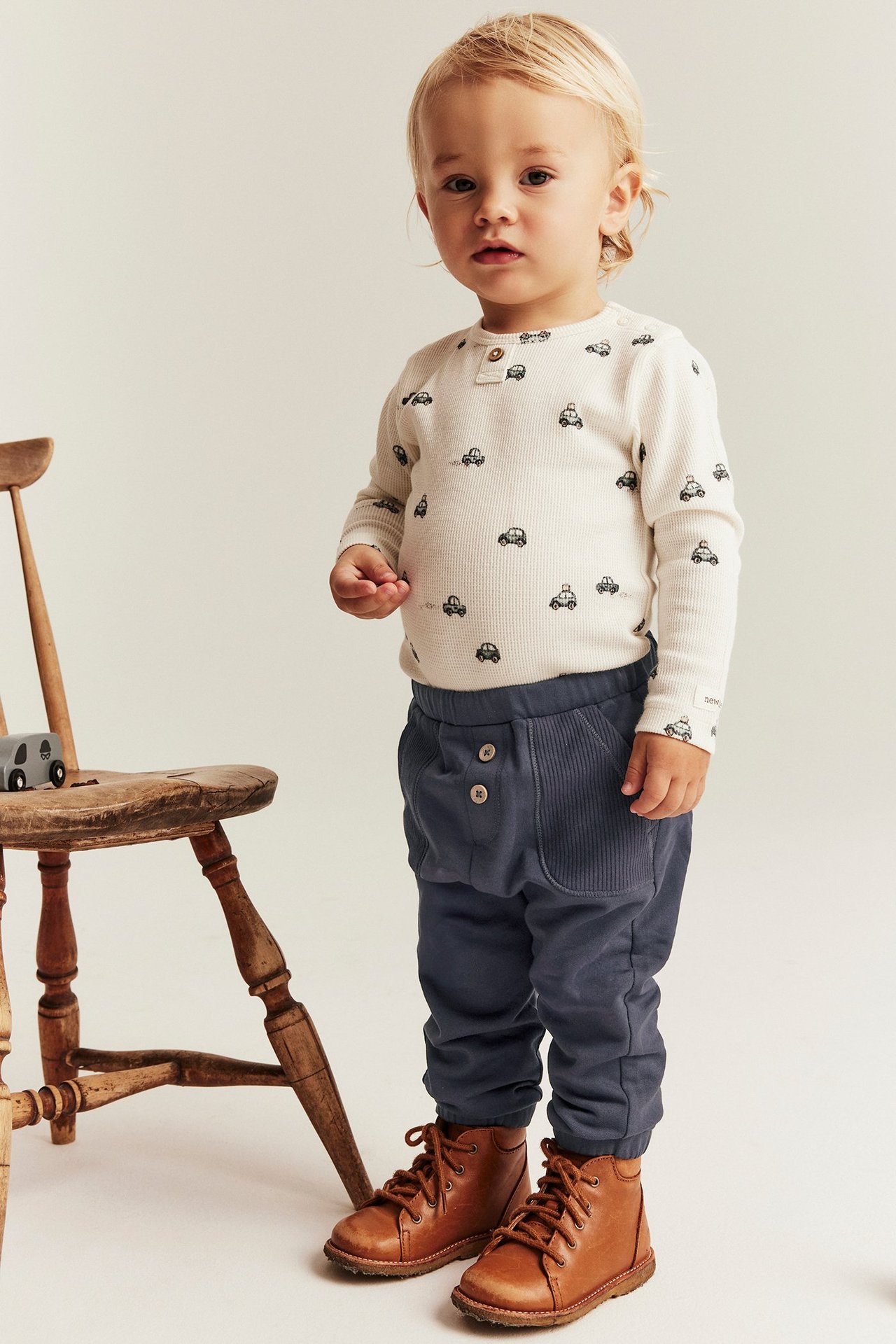 Front view: Kid in dark blue cotton jogging pants, front pockets, decorative buttons, white patterned top, brown boots.