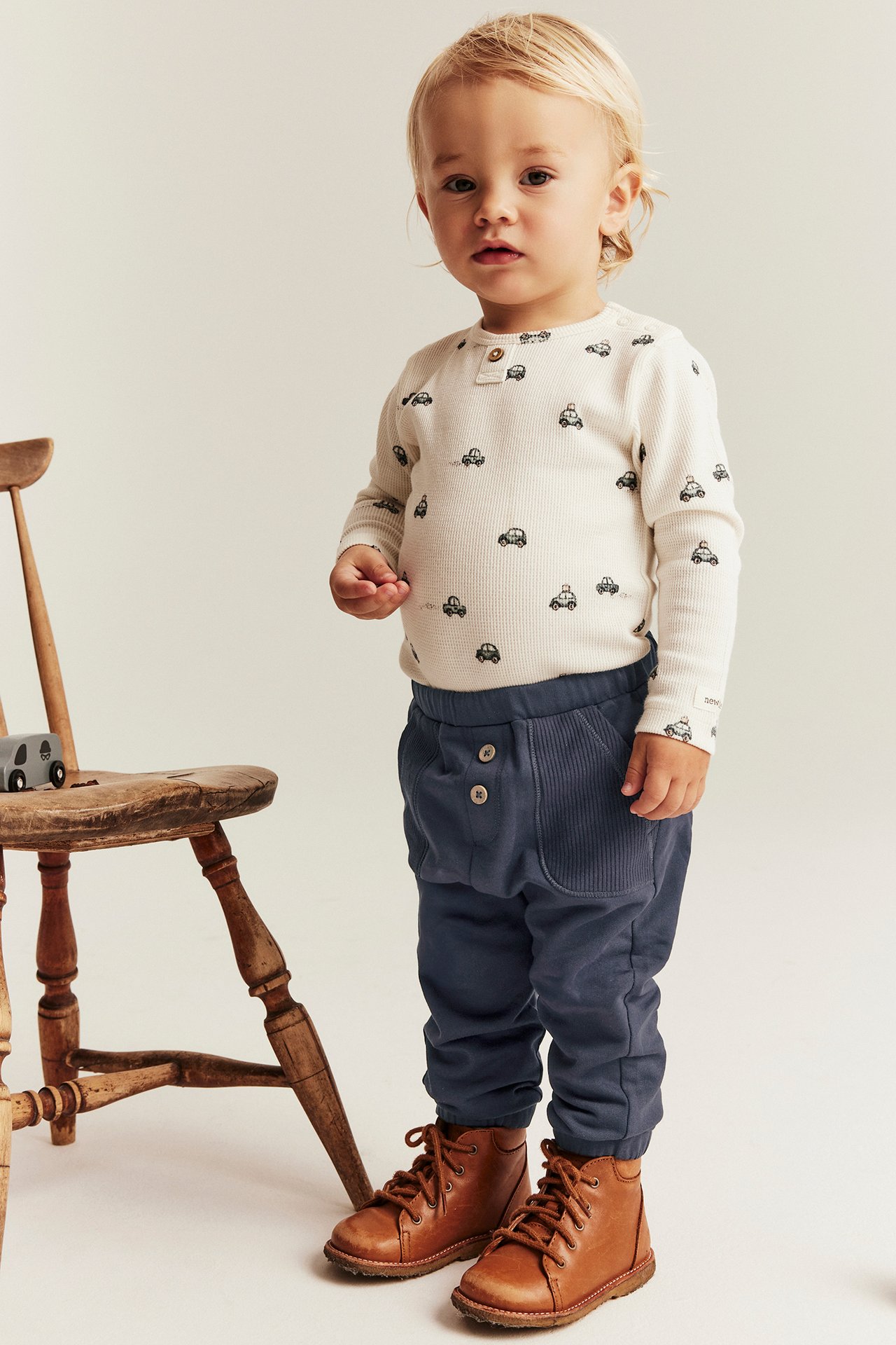Baby trägt dunkelblaue Baumwollhose mit Zierknöpfen, ein weißes Automuster-Oberteil und braune Stiefel.