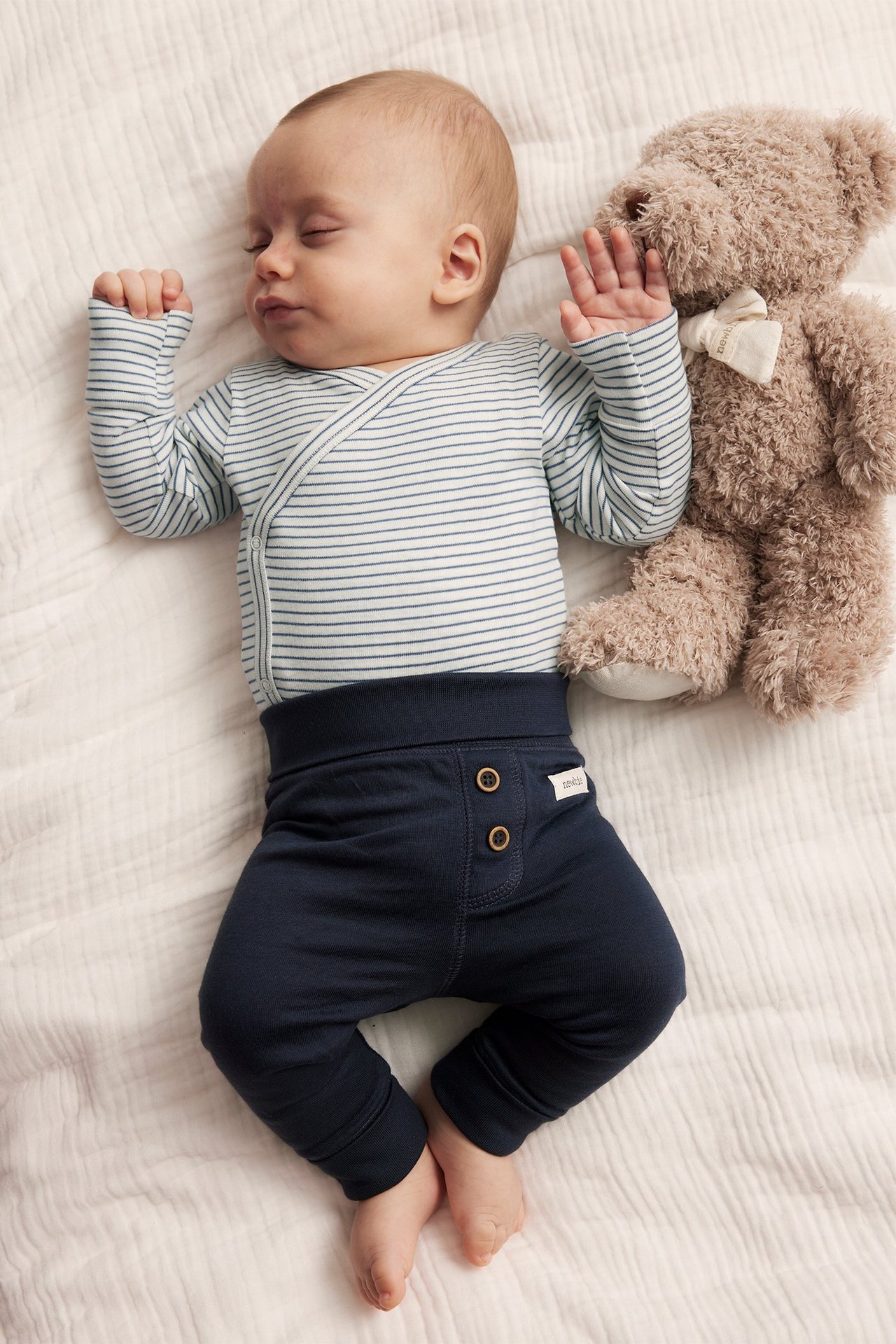 Sleeping baby in a blue striped wrap bodysuit and dark blue pants, with a brown teddy bear.