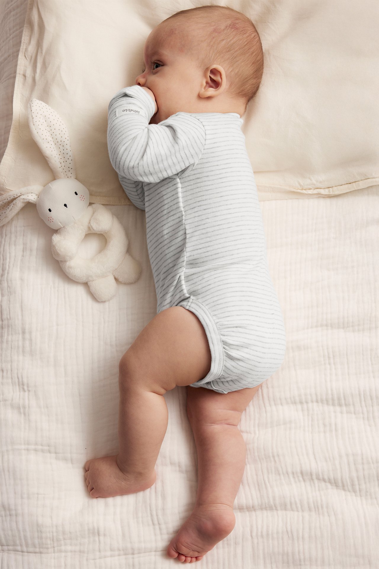 Top-down view: Baby in a grey melange striped long-sleeved wrap bodysuit, next to a white bunny rattle.