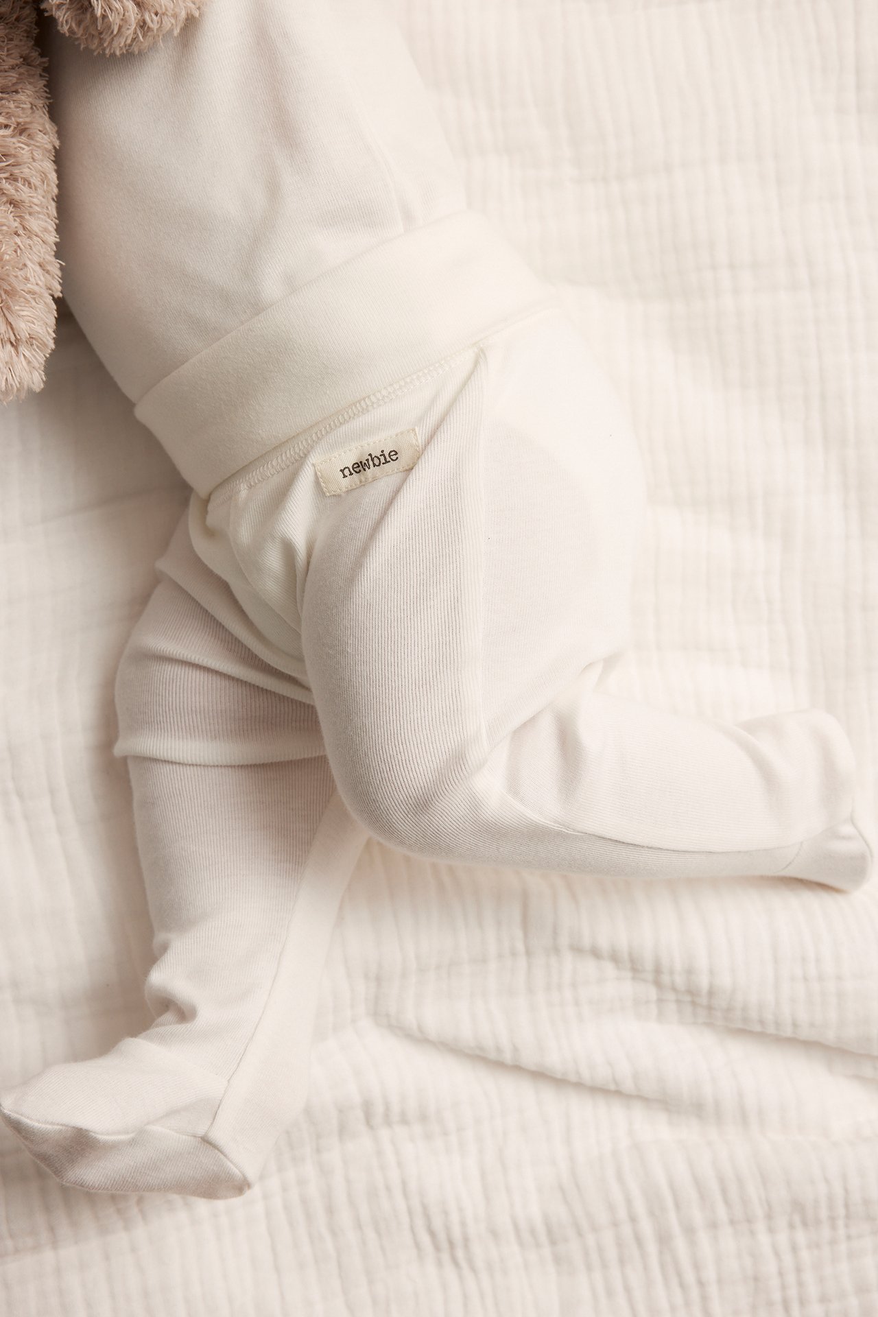 Close-up of baby in off-white cotton footed leggings with wide waist cuff, next to a plush toy.