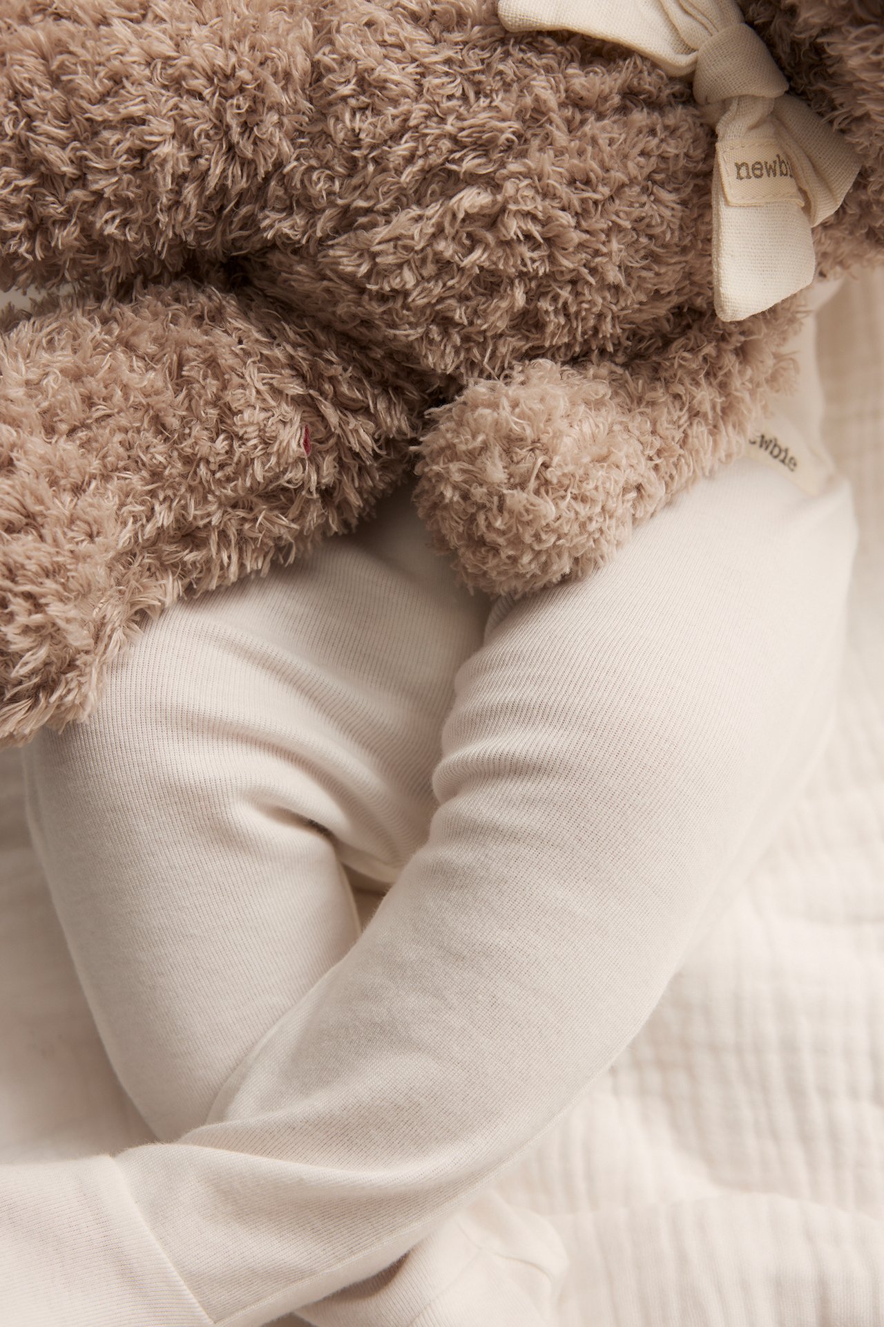 Close-up of a baby wearing offwhite cotton leggings with foot, holding a brown teddy bear.
