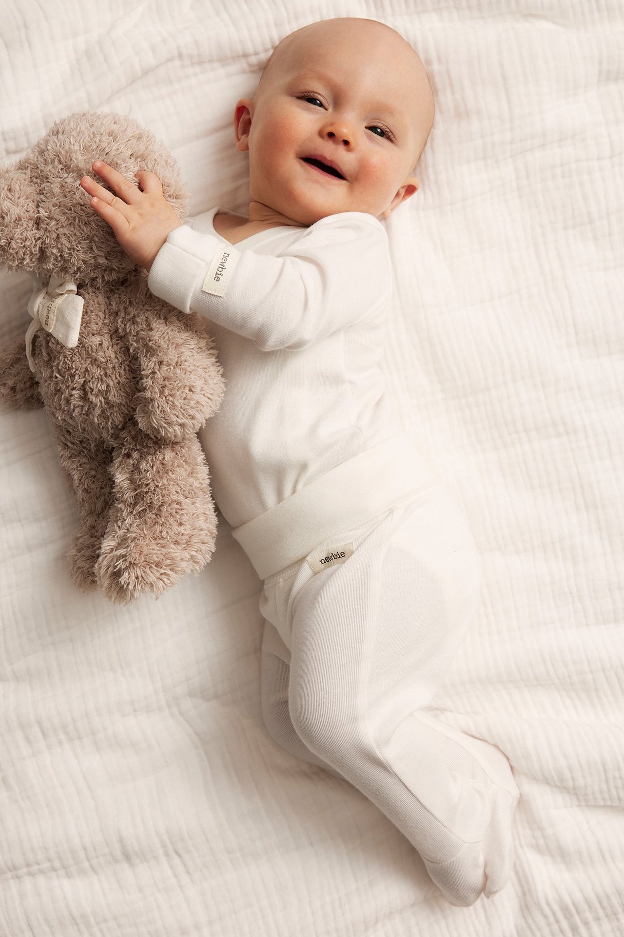 Baby in off-white cotton footed leggings, wide waist, matching top, holding a brown teddy bear.
