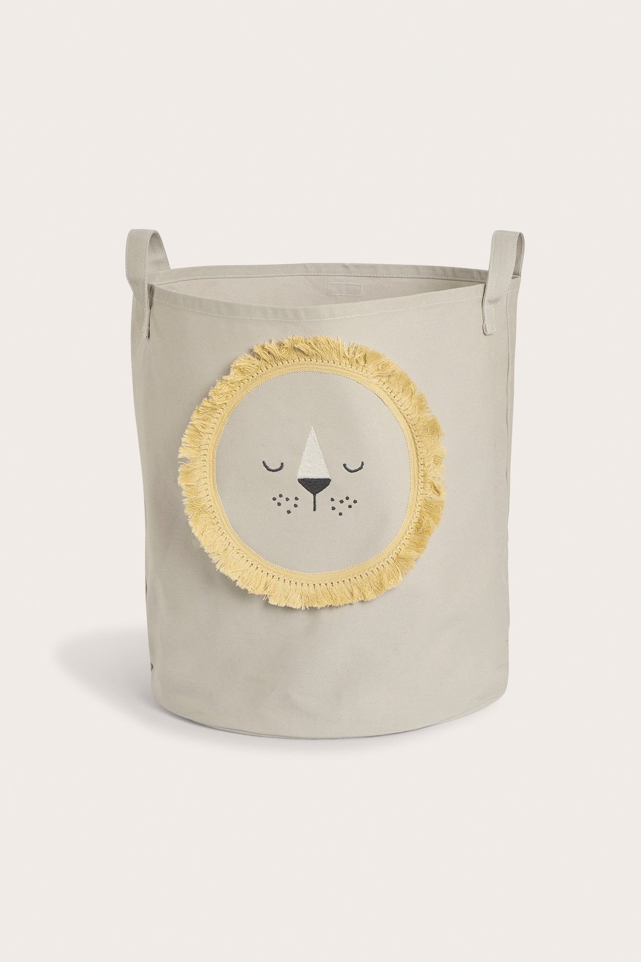 Vorderansicht eines beigen Baumwoll-Aufbewahrungskorbs mit Griffen und Löwengesichts-Applikation mit gelben Fransen.