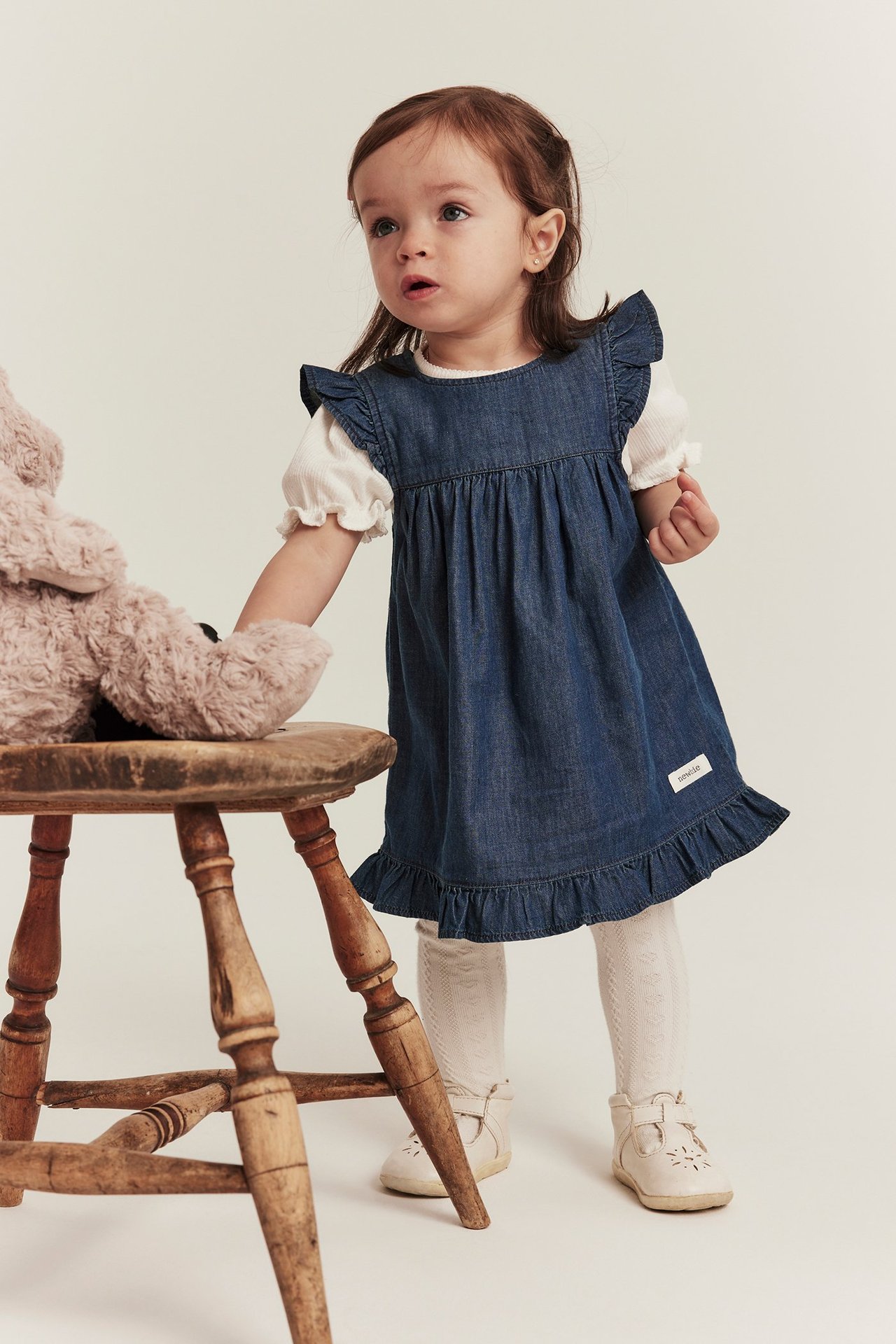 Vorderansicht: Baby trägt ein Jeans-Rüschenkleid über weißem Ripptop, gemusterte Strumpfhosen und beige Schuhe.