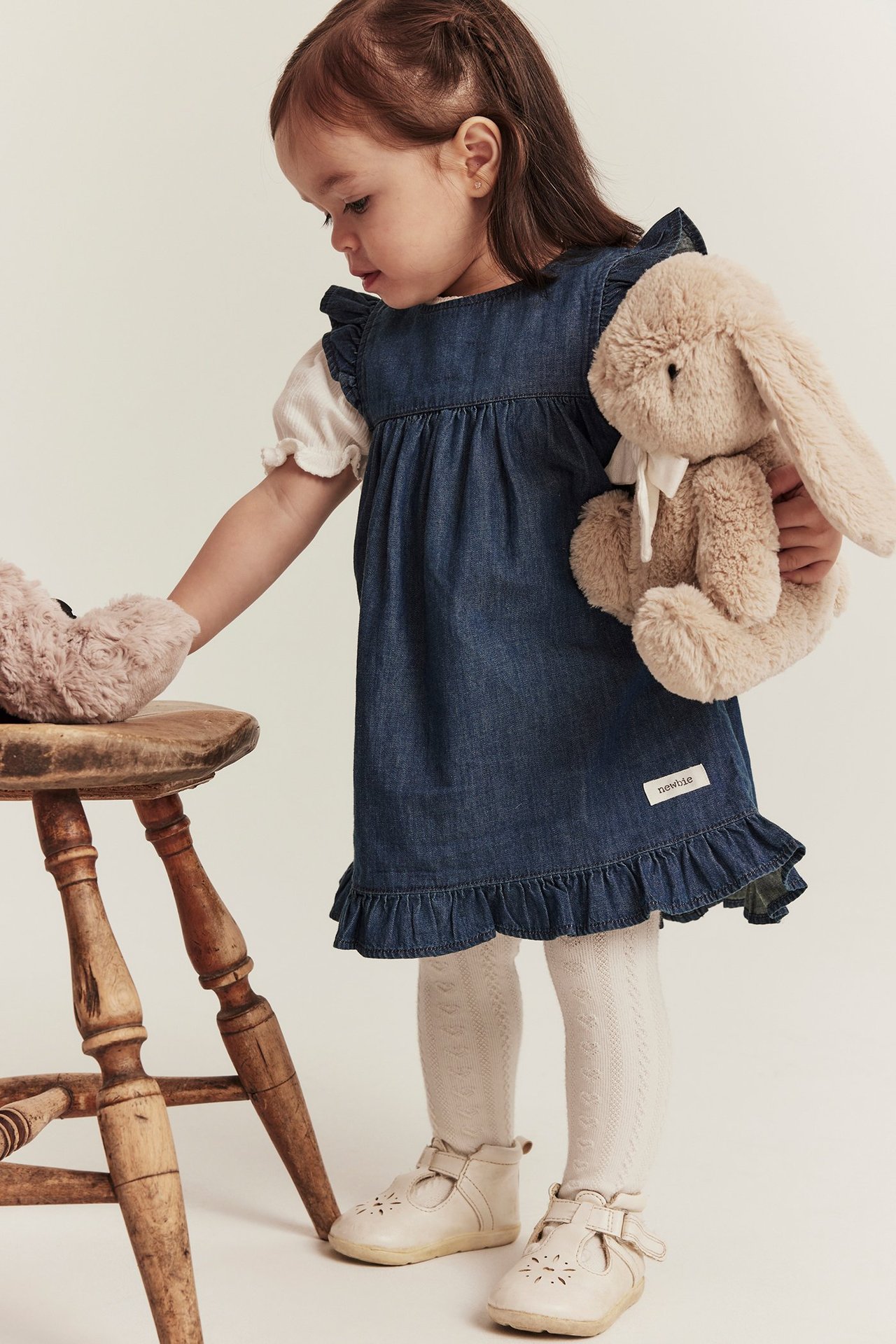 Kind trägt dunkelblaues Jeans-Rüschenkleid, weißes Top, gemusterte Strumpfhose, beige Schuhe, hält braunes Kaninchen.